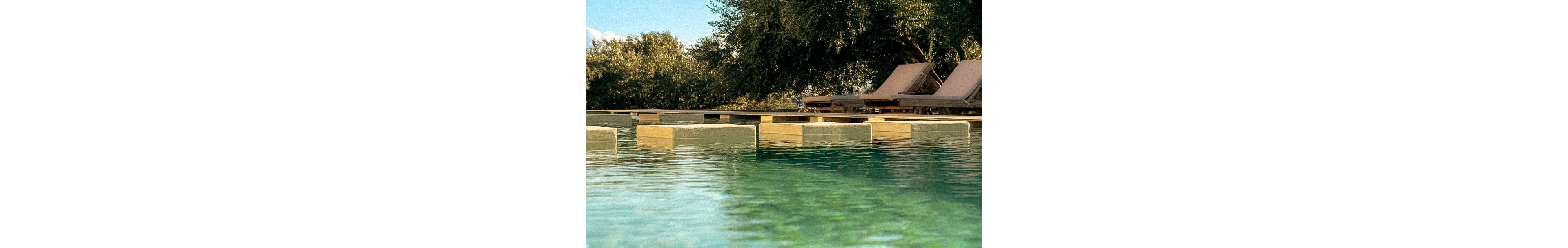 A serene outdoor pool features geometric floating steps leading across the water. In the background, two cushioned lounge chairs are positioned under the shade of lush green trees. The scene is bathed in warm sunlight, creating a tranquil and inviting atmosphere. The water reflects the surrounding greenery, enhancing the relaxing mood.