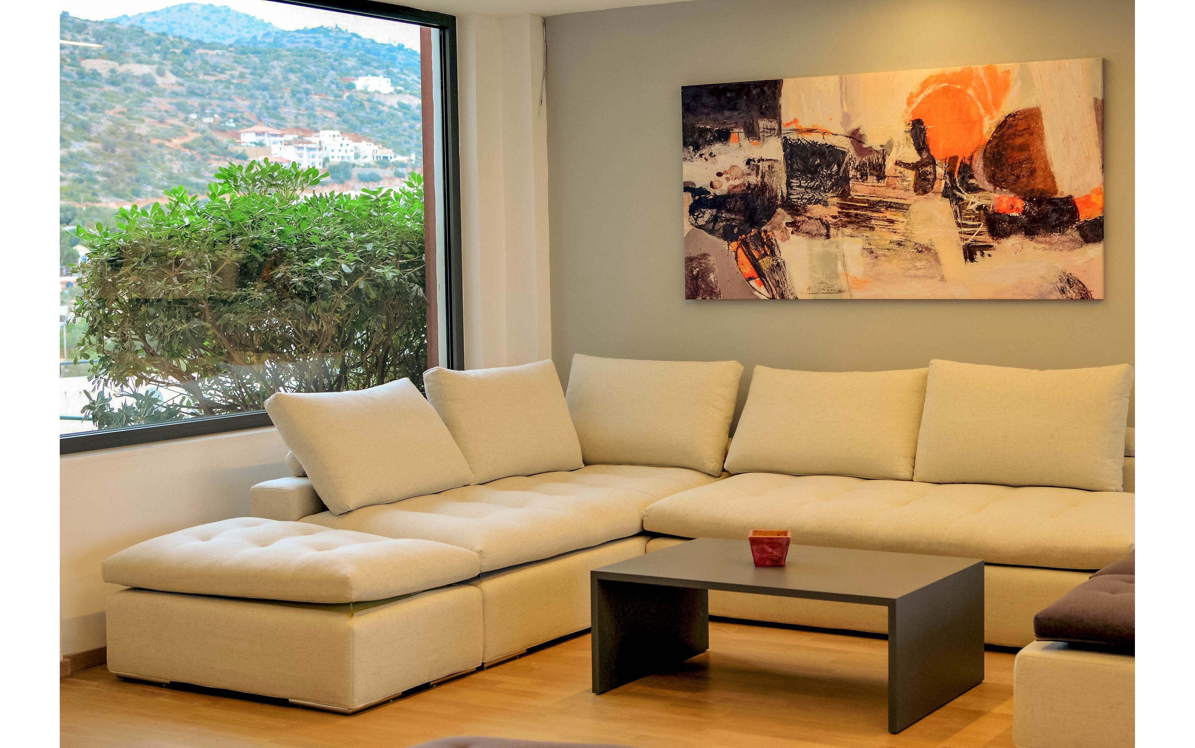 A contemporary living room features a large beige sectional sofa arranged around a low black coffee table. A colorful abstract painting hangs on the wall, adding visual interest to the neutral palette. Natural light streams in from a large window, revealing greenery and a scenic hillside view outside. The space is uncluttered, with a single decorative item on the table, creating a calm and inviting atmosphere.