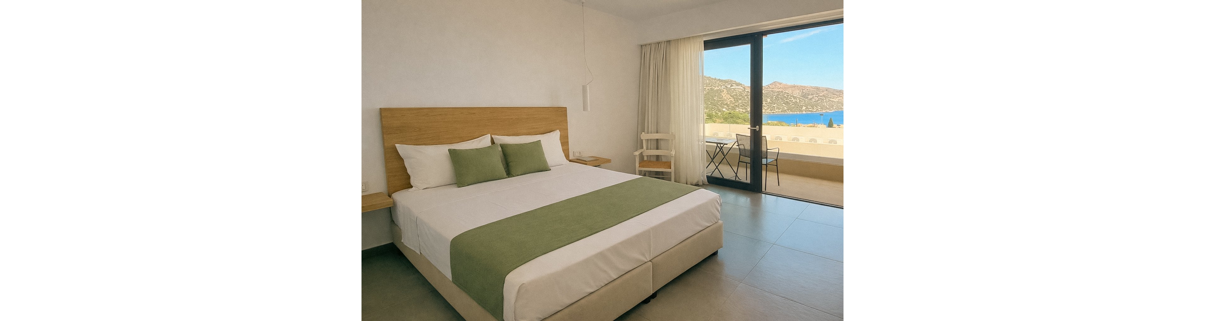 A spacious hotel room features a large bed with green accents and minimalist decor. The room opens to a balcony with a table and chairs, offering a scenic view of the sea and surrounding hills. Natural light fills the space through large glass doors, creating a calm and inviting atmosphere. The setting is clean and uncluttered, ideal for relaxation or vacation stays.