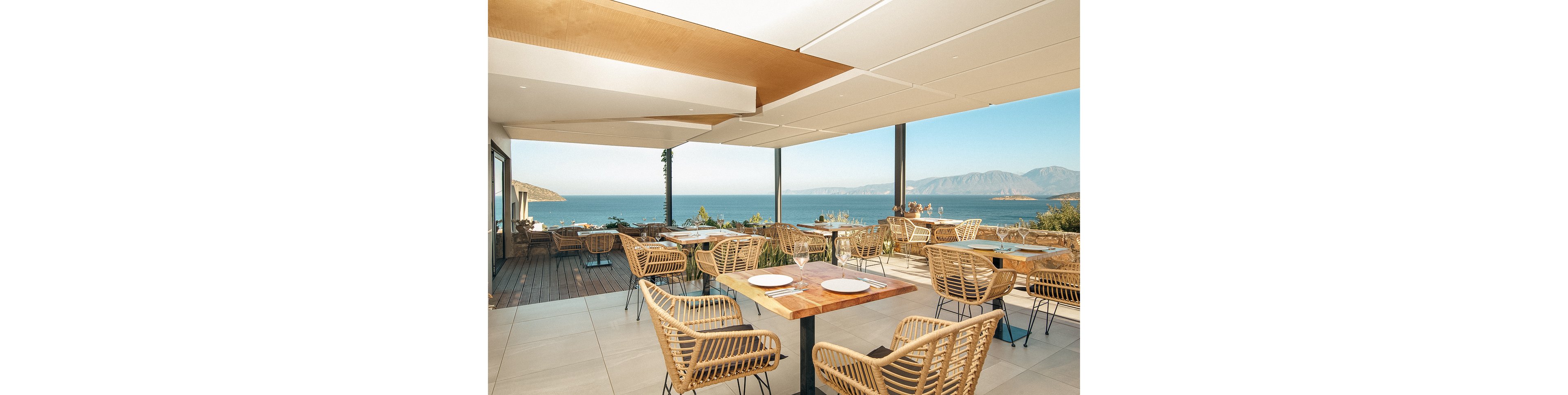 A contemporary open-air restaurant terrace overlooks a calm blue sea and distant mountains. The setting features wooden tables, wicker chairs, and minimalist table settings, creating a relaxed and inviting atmosphere. Natural light floods the space, highlighting the neutral color palette and scenic outdoor environment. No visible people or text are present in the image.