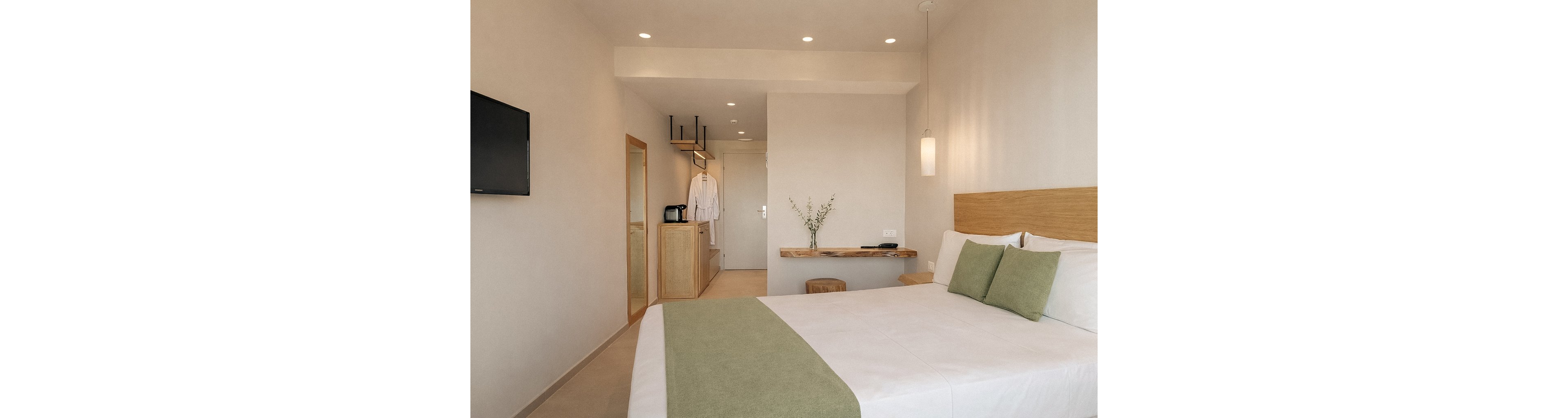 A bright, minimalist hotel room features a neatly made bed with white linens and green pillows. The space includes a wall-mounted TV, a wooden desk with a vase of greenery, and a small kitchenette area with a coffee maker and hanging robes. Soft lighting and neutral tones create a calm, inviting atmosphere, while the uncluttered design emphasizes relaxation and comfort.