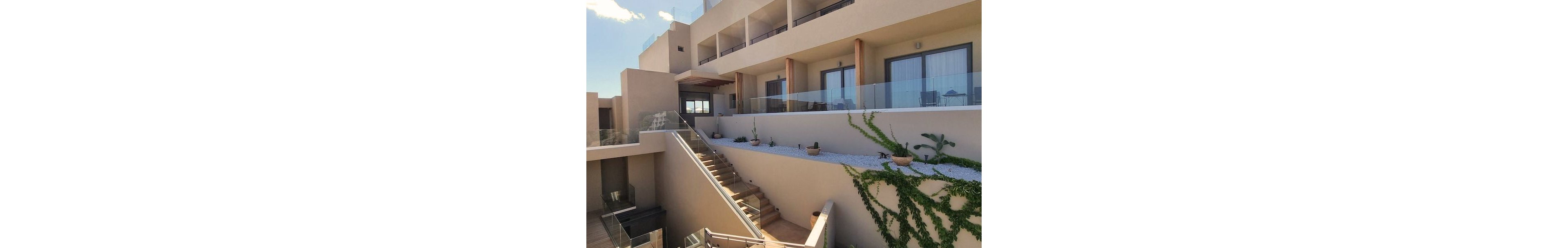 A contemporary hotel building features multiple floors with glass balcony railings and wide outdoor staircases. The setting is bright and sunny, with potted plants and climbing greenery adding a touch of nature to the minimalist architecture. The neutral color palette and clean lines create a calm, inviting atmosphere. No visible text or numbers are present in the image.