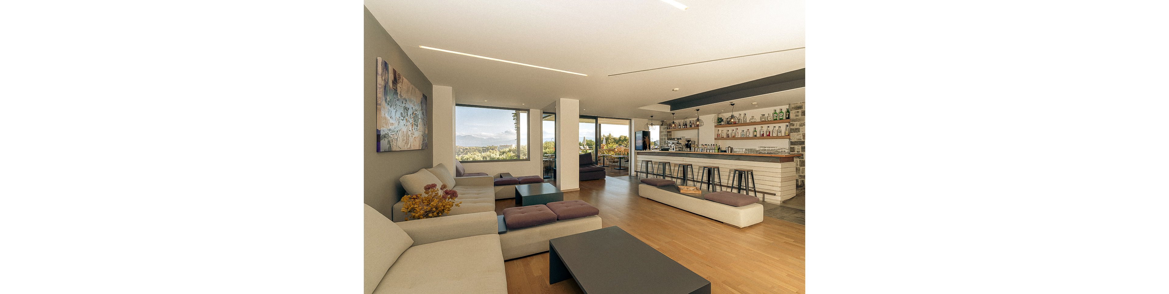 A spacious, contemporary lounge bar features minimalist sofas and a sleek bar counter with high stools. Large windows offer scenic views of greenery and mountains, filling the room with natural light. The interior uses neutral tones with subtle pops of color from decor and wall art. Bottles and glassware are neatly arranged on shelves behind the bar. The atmosphere is relaxed and inviting, ideal for social gatherings or casual meetings.