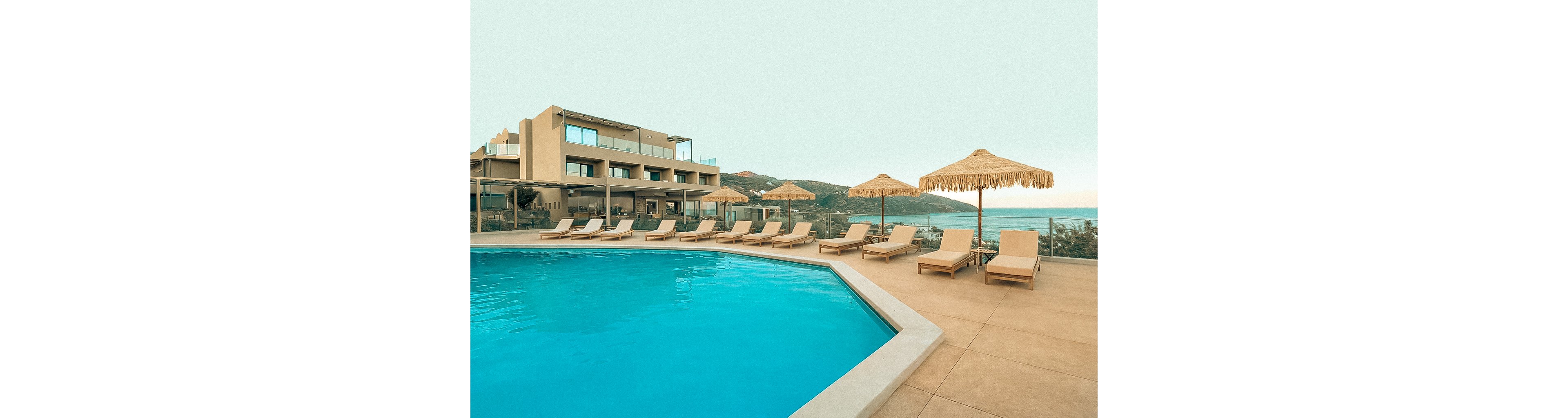 A contemporary resort features a large outdoor swimming pool surrounded by beige sun loungers and straw parasols. The setting overlooks a scenic coastline with hills in the background, creating a tranquil and inviting atmosphere. The pool area is empty, emphasizing relaxation and leisure in a serene environment. The color palette is warm and neutral, with soft blue water contrasting sandy tones.
