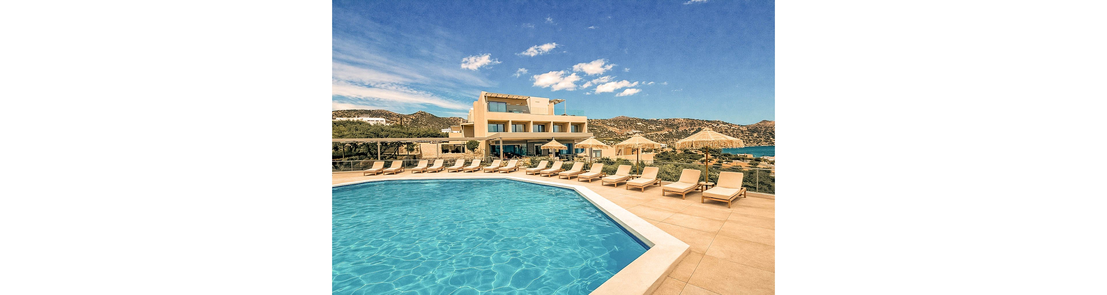 A modern resort features a large outdoor swimming pool surrounded by numerous sun loungers and straw parasols. The setting is bright and sunny, with clear blue skies and scenic hills in the background. The architecture is contemporary, and the overall mood is relaxed and inviting, ideal for vacation or leisure. No visible text or numbers are present in the image.