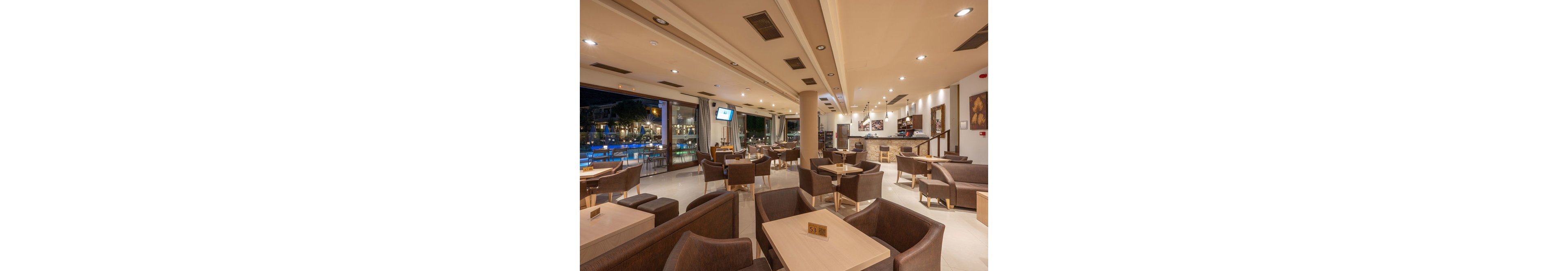 The image shows a spacious hotel lounge featuring contemporary brown chairs and light wood tables. The setting is indoors with large windows overlooking a lit swimming pool area at night. The mood is relaxed and inviting, with warm neutral tones and soft lighting. Table numbers such as '53' are visible, and a television screen is mounted on the wall. The bar area is seen in the background, adding to the social ambiance.