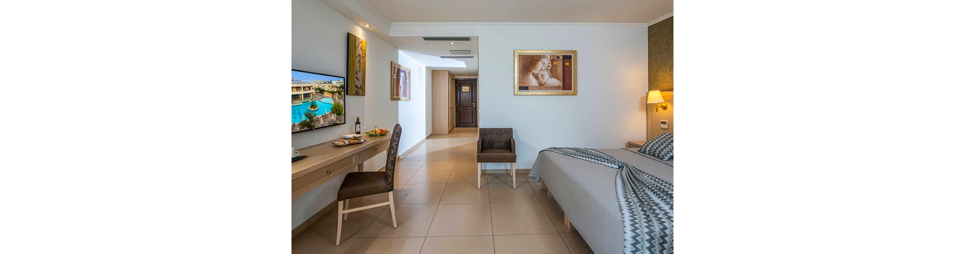 A contemporary hotel room features a neatly made bed, a wall-mounted television displaying a scenic landscape, and a desk with a chair and food items. The space is decorated with framed artwork and neutral tones, creating a calm and inviting atmosphere. The room is well-lit, with tiled flooring and a hallway leading to the entrance.
