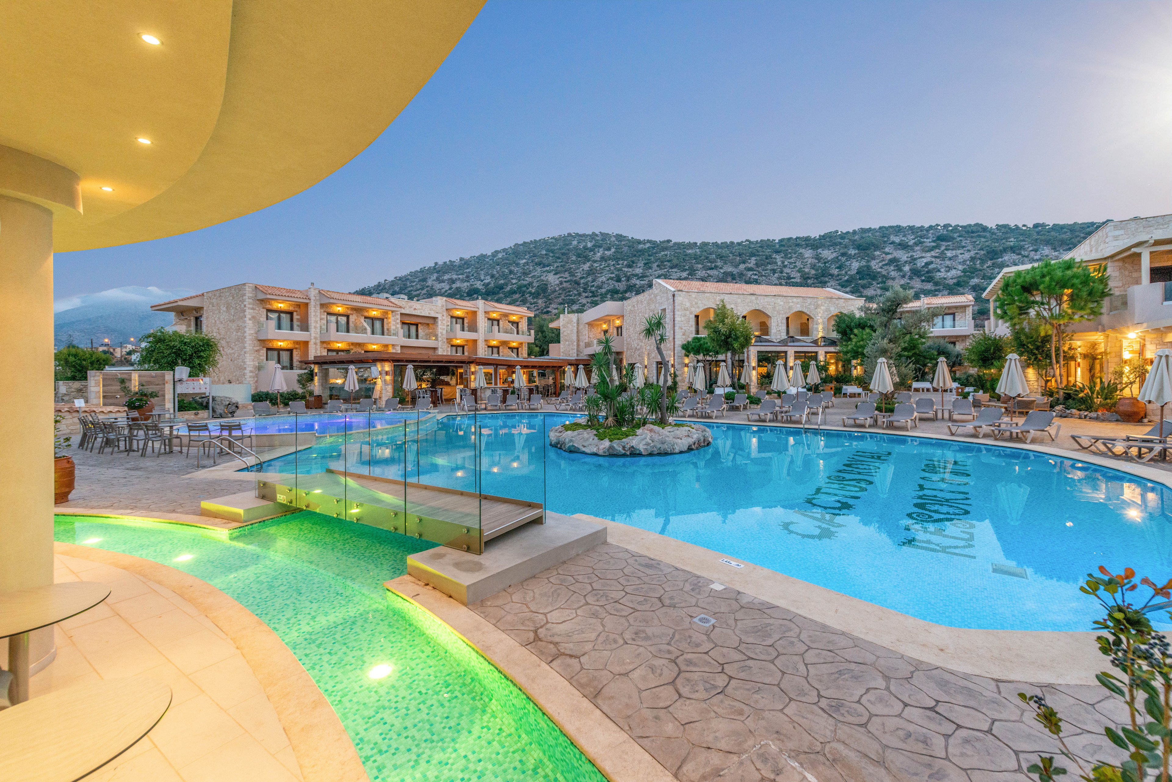 A spacious outdoor swimming pool is the focal point of this upscale resort, surrounded by lounge chairs and modern stone buildings. The tranquil setting features soft evening lighting, clear blue water, and a backdrop of rolling hills. The scene exudes a relaxing and inviting atmosphere, ideal for leisure and vacation stays.