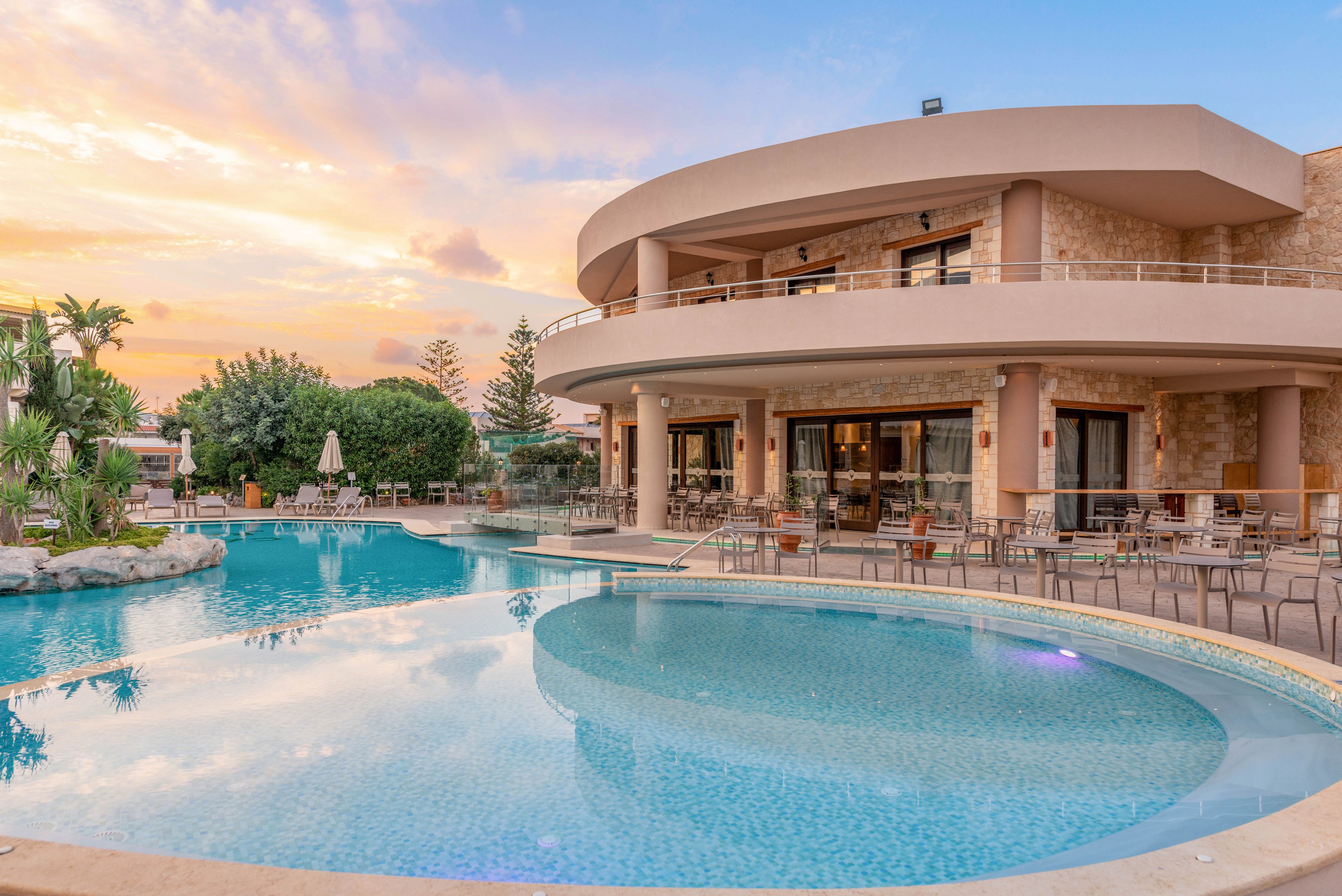 A contemporary resort building features a large, curved outdoor swimming pool surrounded by lounge chairs and tables. The setting is tranquil, with palm trees and landscaped greenery enhancing the relaxing atmosphere. The sky is painted with soft pastel hues as the sun sets, reflecting off the calm water. The architecture is modern with stone and glass elements, creating an inviting vacation ambiance.