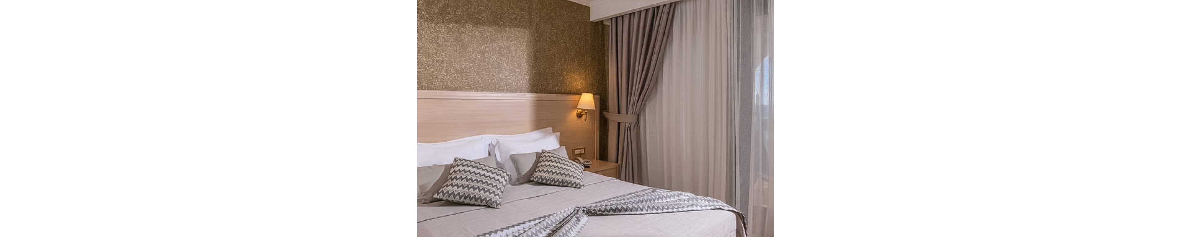 A contemporary hotel room features a large double bed with white linens and chevron-patterned pillows and throw. The room is softly lit by a bedside lamp mounted on a textured gold accent wall. Floor-to-ceiling curtains frame a large window, allowing natural light to filter in, and a small nightstand sits beside the bed.