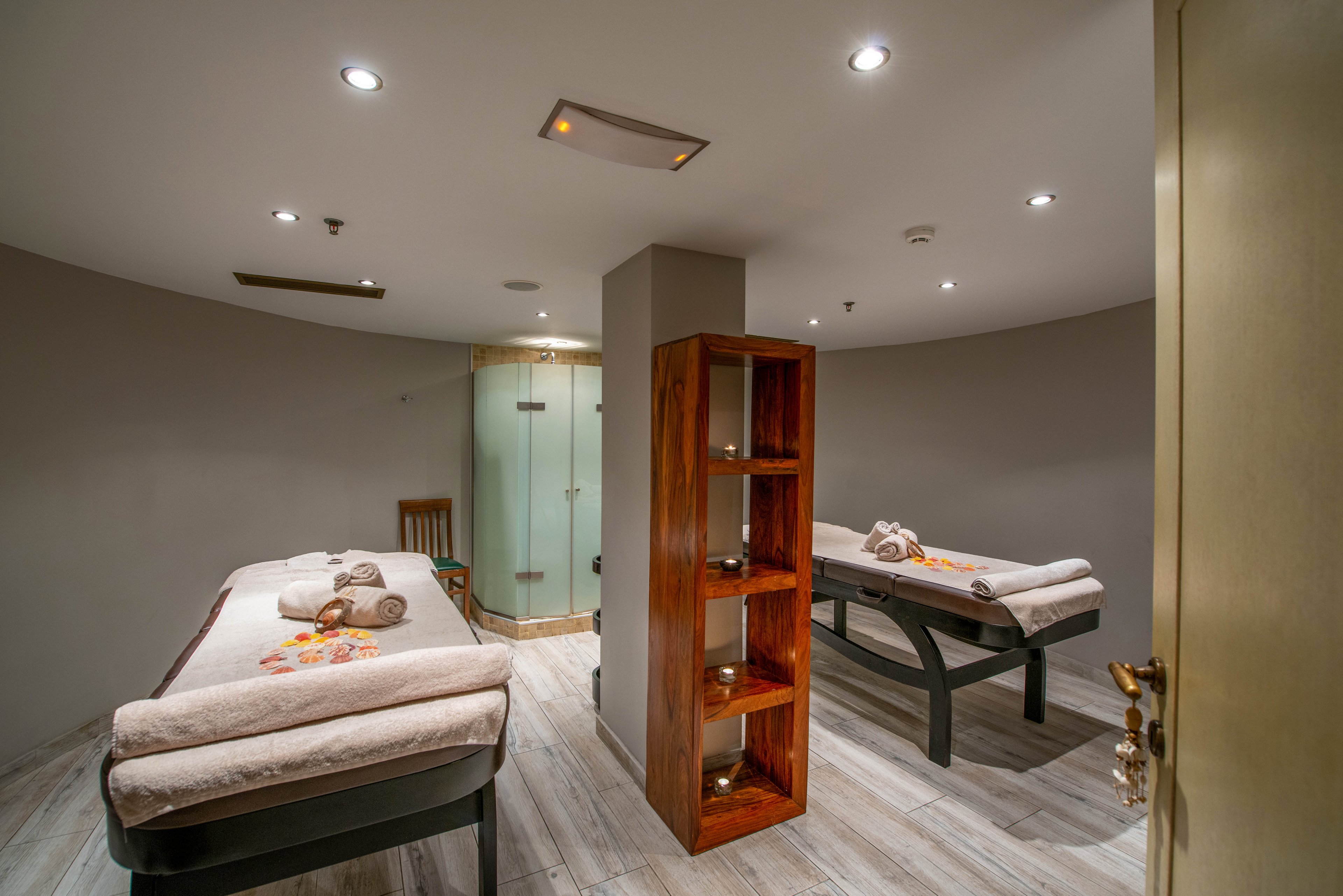 A contemporary spa treatment room features two massage tables neatly arranged with rolled towels and flower petals. The space is softly lit with recessed ceiling lights, creating a calm and relaxing atmosphere. A wooden shelving unit divides the room, and a glass shower enclosure is visible in the background. The neutral color palette and minimalist decor enhance the tranquil mood.