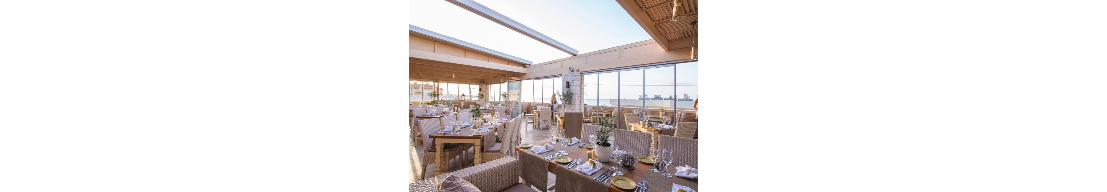 A bright, airy rooftop restaurant features neatly arranged tables set for dining, each adorned with white napkins, glassware, and potted plants. The setting overlooks the ocean, visible through large windows, and is bathed in natural sunlight. The decor is modern with light wood tones and neutral upholstery, creating a relaxed and inviting atmosphere. A few people are seen in the background, enhancing the lively yet calm ambiance.