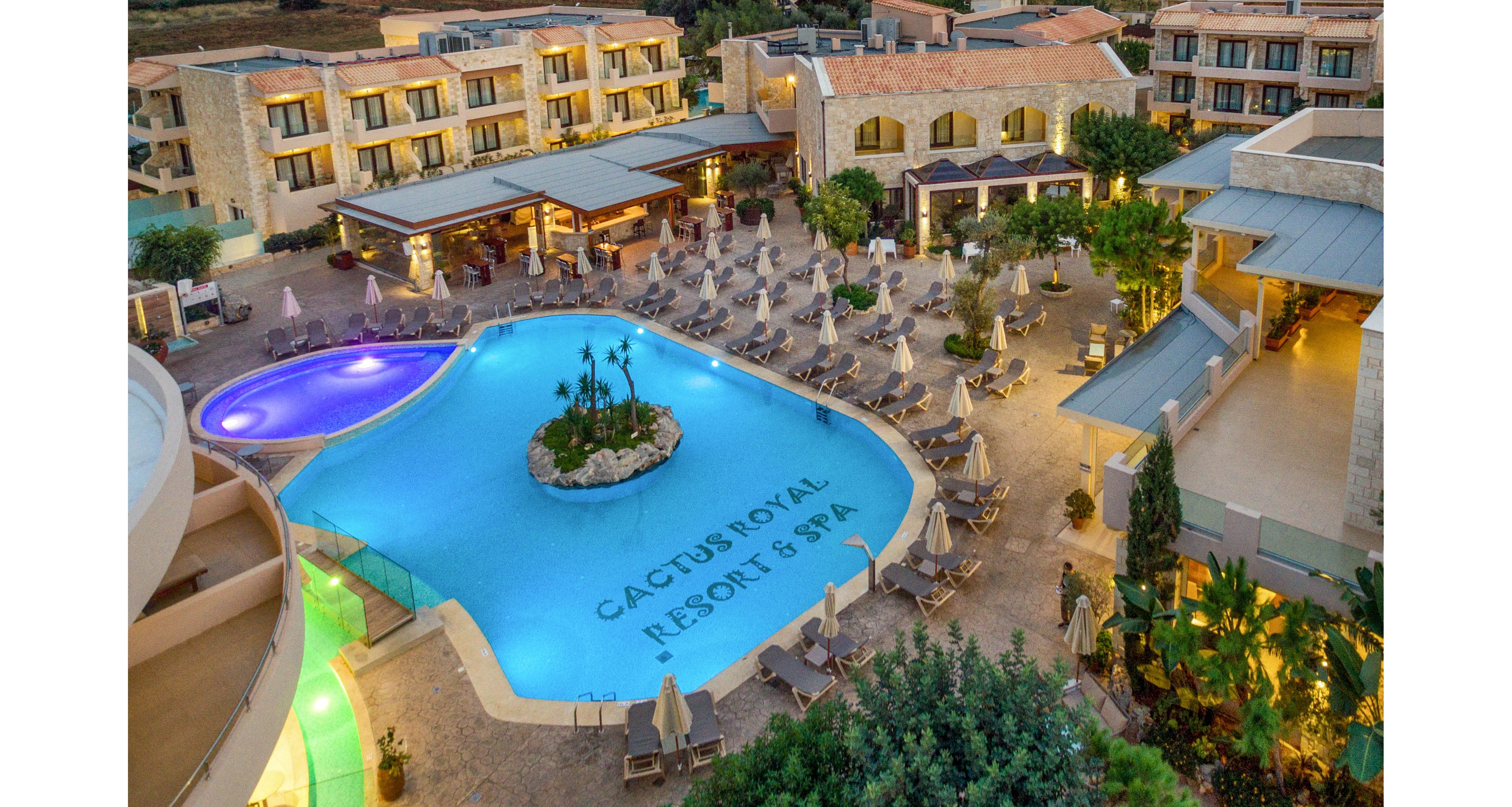This image shows an aerial view of the Cactus Royal Resort & Spa, featuring a large, uniquely shaped swimming pool at the center. The pool area is surrounded by numerous lounge chairs and umbrellas, with landscaped greenery and resort buildings in the background. The setting appears tranquil and inviting, ideal for relaxation and leisure. The text 'CACTUS ROYAL RESORT & SPA' is clearly visible on the pool floor.