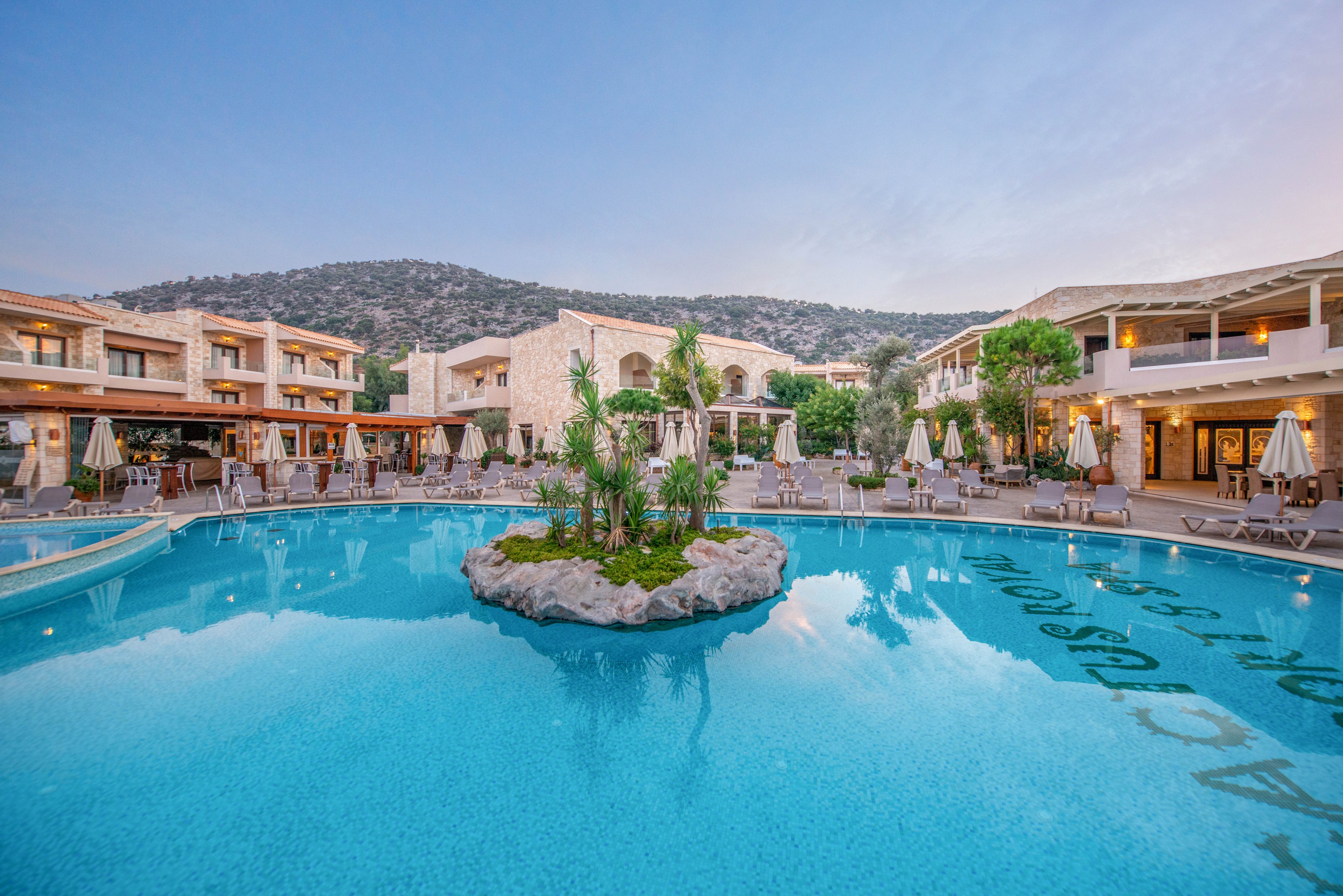 A tranquil resort pool is the central focus, surrounded by sun loungers and umbrellas. The setting features modern stone buildings and lush landscaping, with a small island of greenery in the pool. The scene is set against a backdrop of rolling hills under a clear evening sky, creating a relaxing and inviting atmosphere. No visible text or numbers are present in the image.