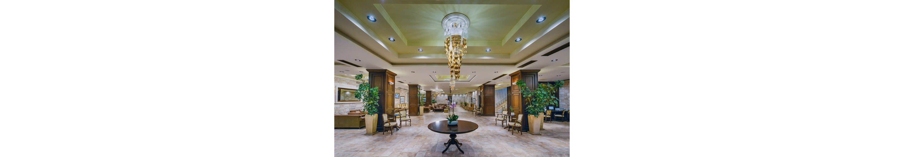 A spacious hotel lobby features a grand chandelier as its centerpiece, hanging above a round table with a decorative orchid. The setting includes plush seating areas, potted plants, and polished tile flooring, creating a welcoming and luxurious atmosphere. Warm lighting and neutral tones contribute to a sophisticated and inviting mood. The lobby is empty, emphasizing the architectural details and interior design.