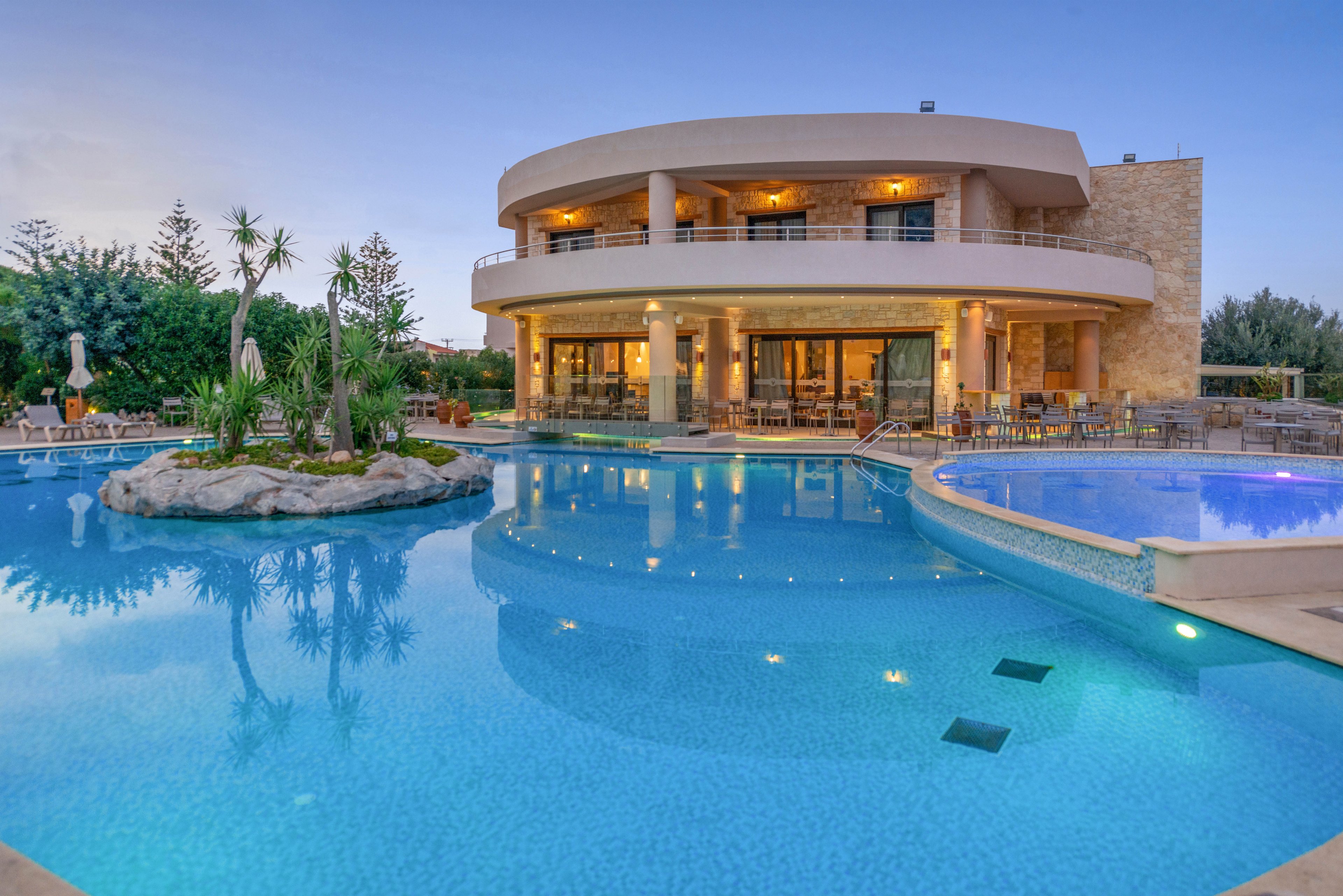A contemporary two-story resort building is featured with large windows and balconies, warmly lit in the early evening. The foreground showcases a spacious, clear blue swimming pool with a small landscaped island and surrounding lounge chairs. The tranquil setting is enhanced by the soft lighting and calm water, creating a relaxing and inviting atmosphere.