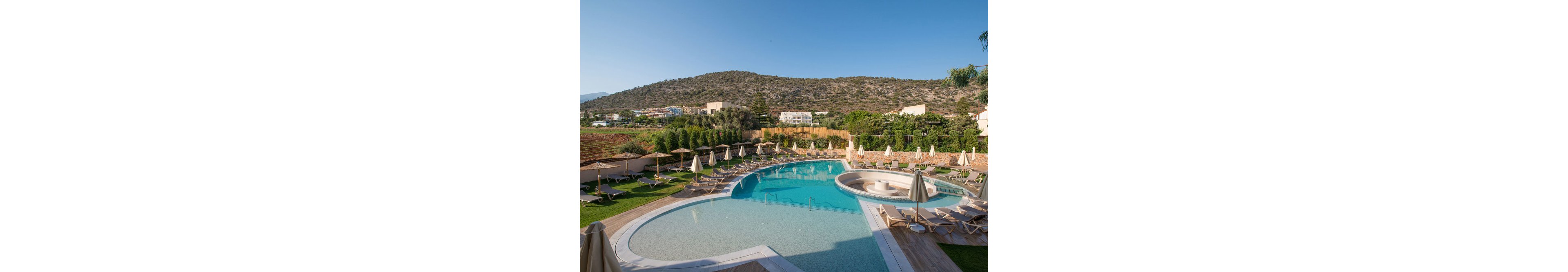 A large, curved outdoor swimming pool is the main focus, surrounded by numerous sun loungers and beige umbrellas. The pool area is set against a backdrop of green hills and scattered buildings, suggesting a resort or hotel setting. The scene is bright and tranquil, with clear blue skies and a relaxed atmosphere. No visible text or numbers are present in the image.