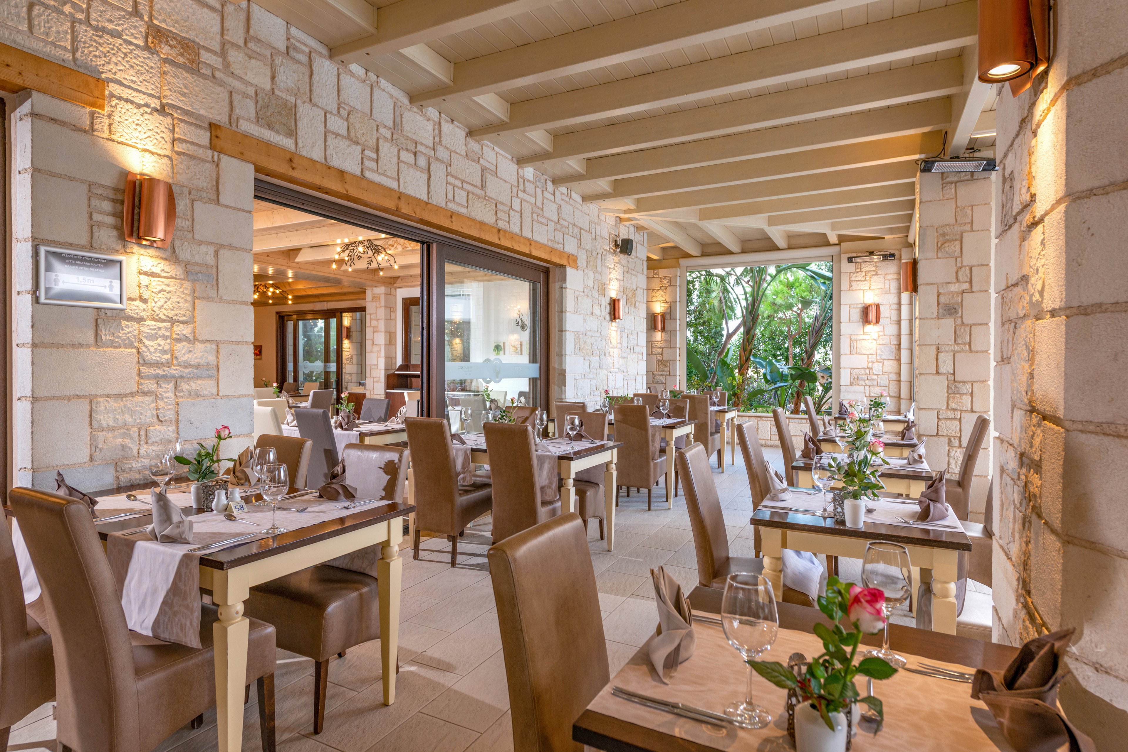 A stylish restaurant dining area features neatly arranged tables with brown leather chairs and white tablecloths. The setting is bright and airy, with natural stone walls and large windows overlooking greenery. Table settings include wine glasses, napkins, and small floral arrangements, creating a welcoming and refined atmosphere. The palette is neutral with warm beige and brown tones, and the space is empty of people.