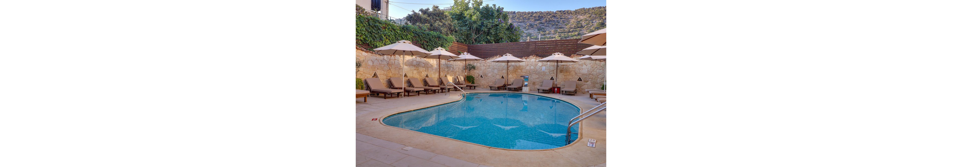 A tranquil outdoor swimming pool is surrounded by cushioned sun loungers and large beige umbrellas. The pool area is enclosed by a stone wall and lush greenery, with hills visible in the background. The clear blue water reflects the serene atmosphere, and poolside numbers such as '1.10' and '1.40' are visible on the tiles, indicating depth in meters.
