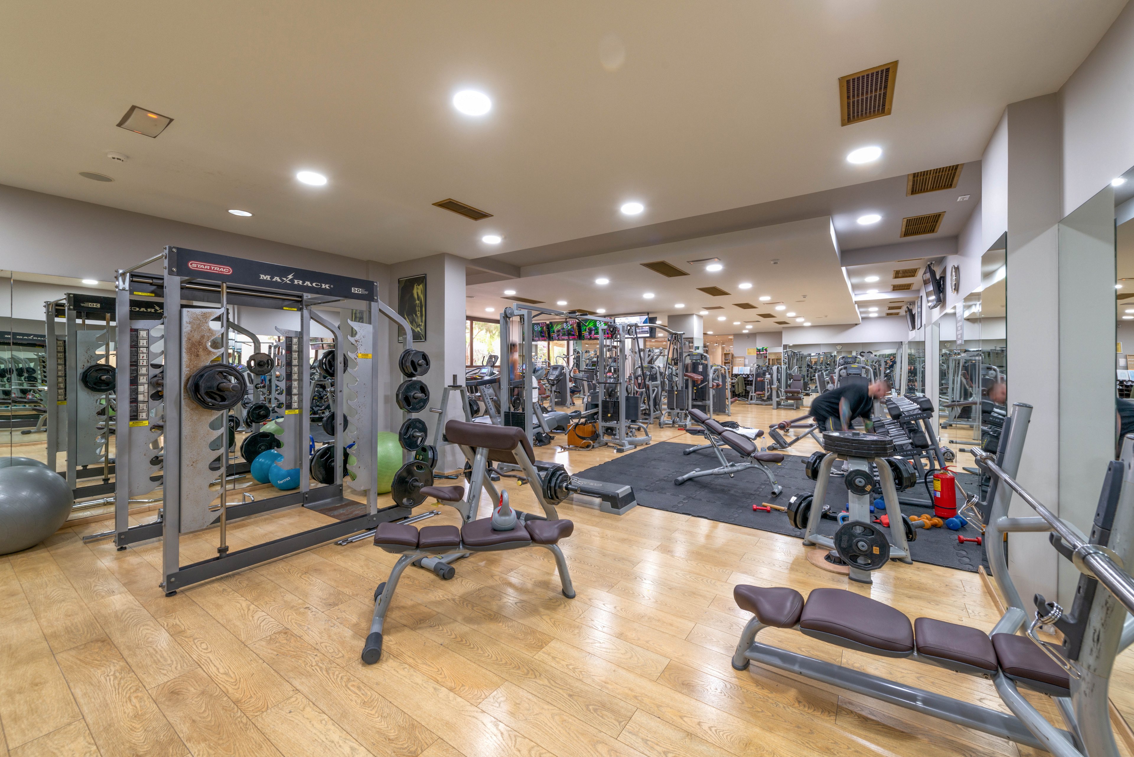 A spacious gym features various weight machines, benches, and free weights arranged across a polished wooden floor. Several adults are engaged in strength training activities, surrounded by mirrors and bright overhead lighting. The setting is clean and organized, with a neutral color palette and a focus on fitness equipment. No visible text or numeric values are present on the machines or walls.