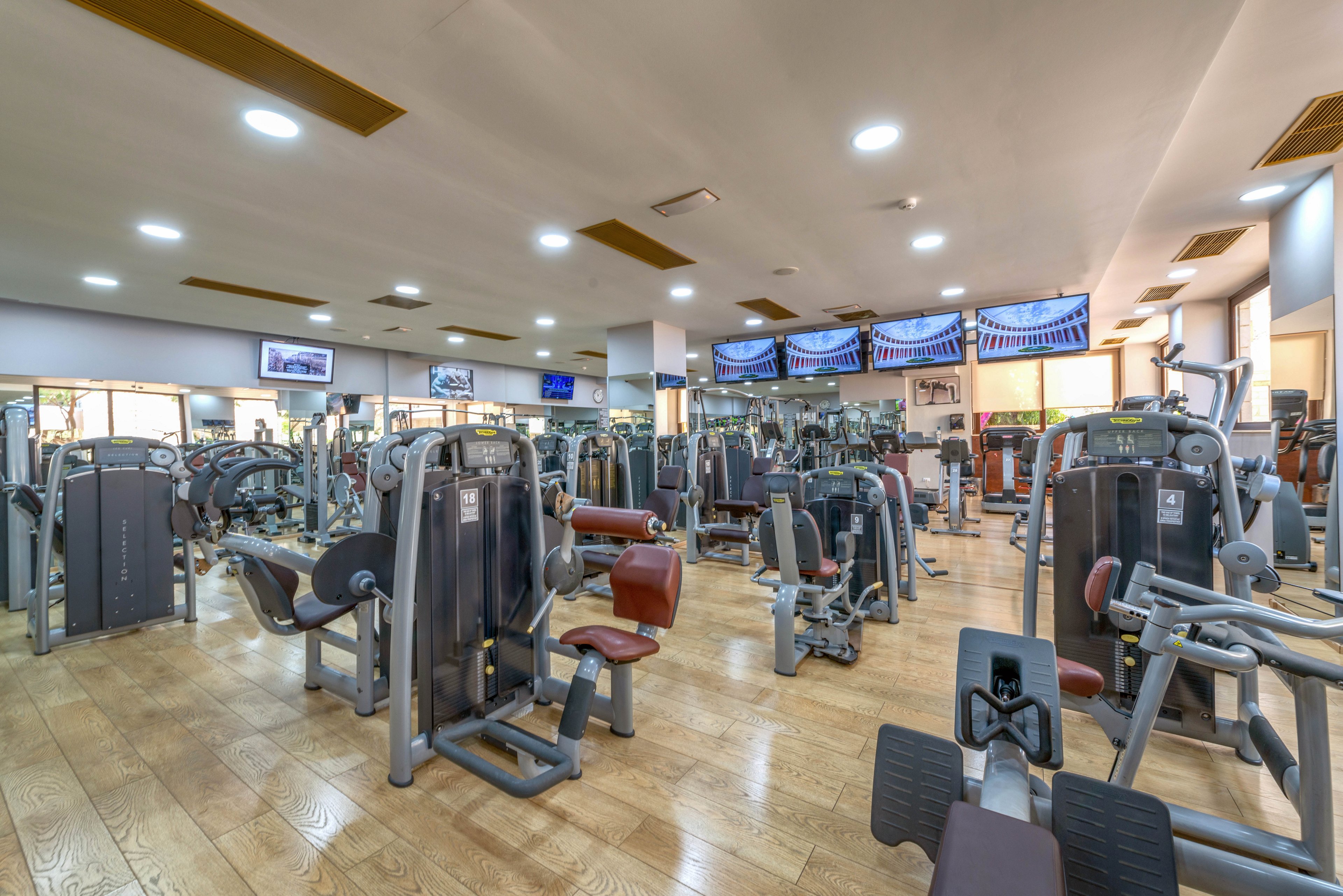 A spacious gym interior featuring a variety of exercise machines arranged neatly across a wooden floor. Multiple digital screens are mounted on the walls, displaying visuals above the workout area. The setting is brightly lit with overhead lights, creating a clean and energetic atmosphere suitable for fitness activities.