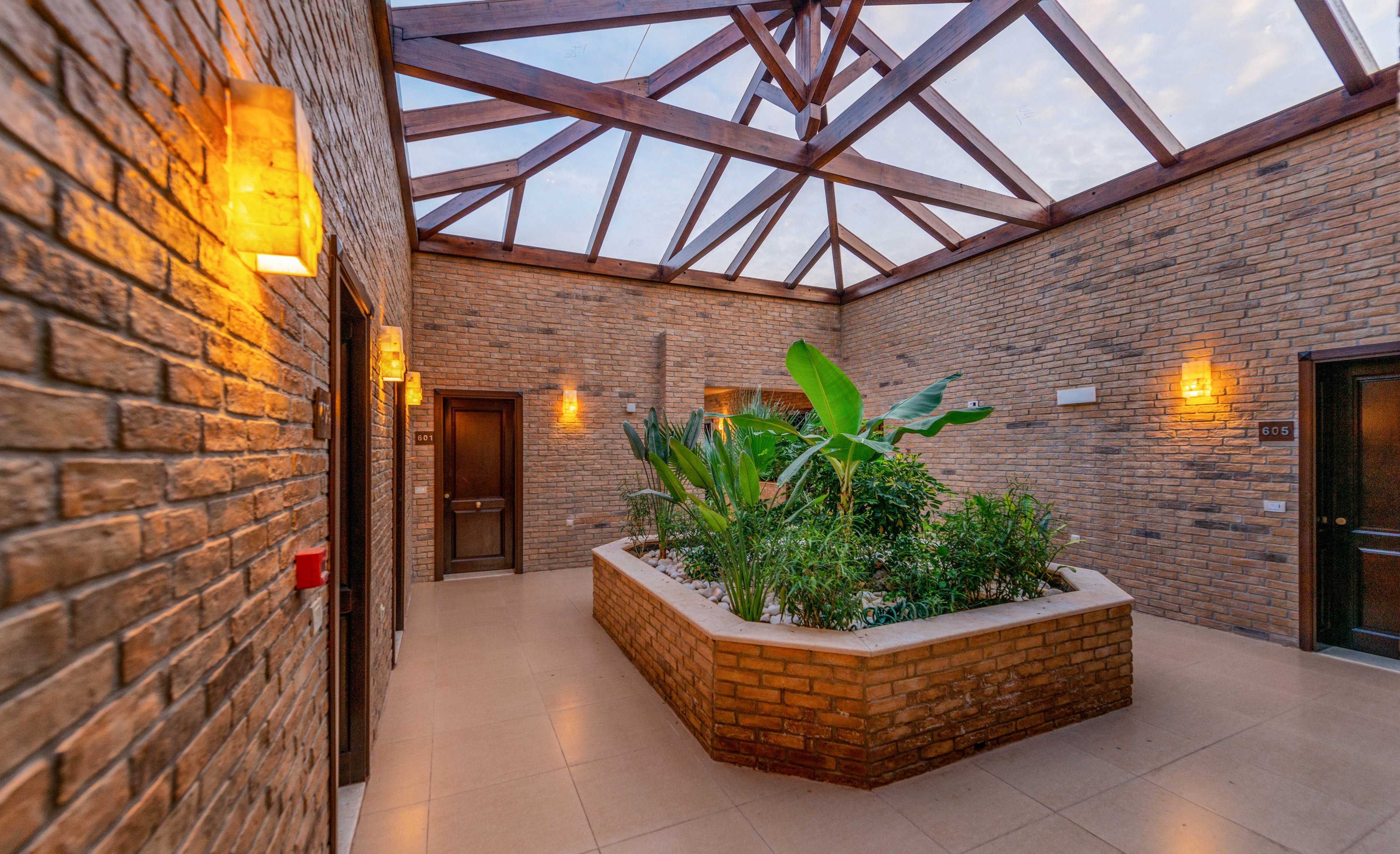 A contemporary indoor atrium features a central planter filled with lush green plants beneath a geometric glass skylight. The space is enclosed by exposed brick walls and illuminated by warm wall sconces. Multiple doors lead off the atrium, and the clean tile flooring enhances the modern aesthetic. No visible text or numbers are present in the image.