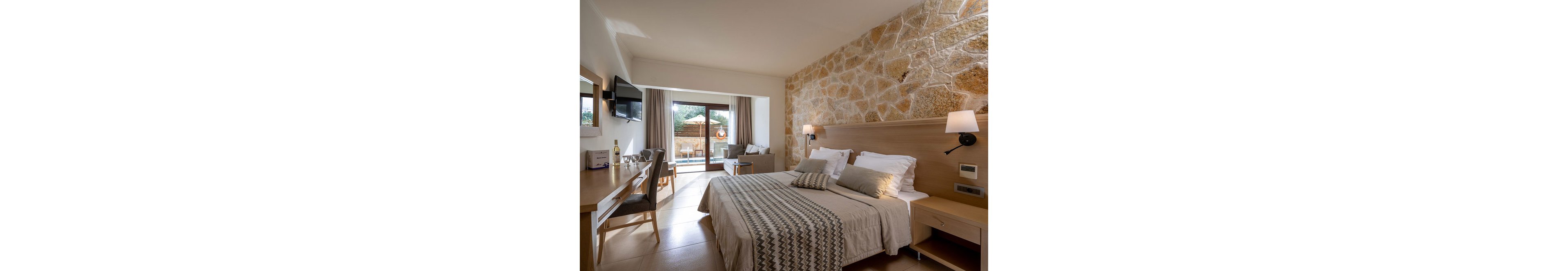 A contemporary hotel room features a double bed with patterned linens, a wooden desk, and a cozy seating area. The space is highlighted by a textured stone accent wall and neutral color palette, creating a warm and inviting atmosphere. Large glass doors open to a private patio with outdoor seating and a pool, allowing natural light to fill the room. No visible text or numbers are present in the image.
