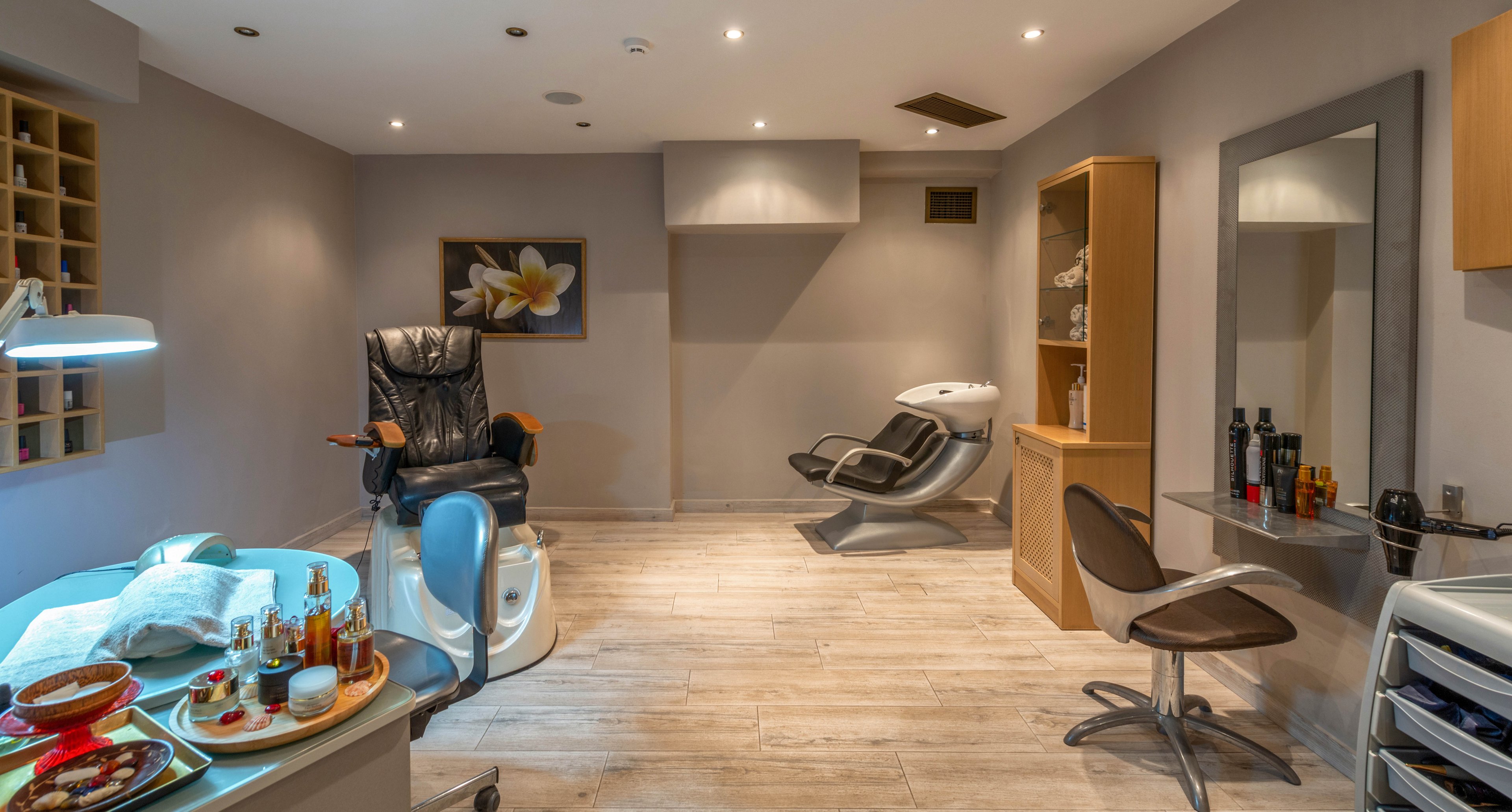 A contemporary spa treatment room features a black massage chair and a white pedicure station. The setting includes a manicure table with beauty products, a styling chair, and a wash basin. The space is softly lit with recessed ceiling lights, neutral walls, and wooden flooring, creating a calm and inviting atmosphere. Decorative elements include a framed flower artwork and neatly arranged bottles on the counter.