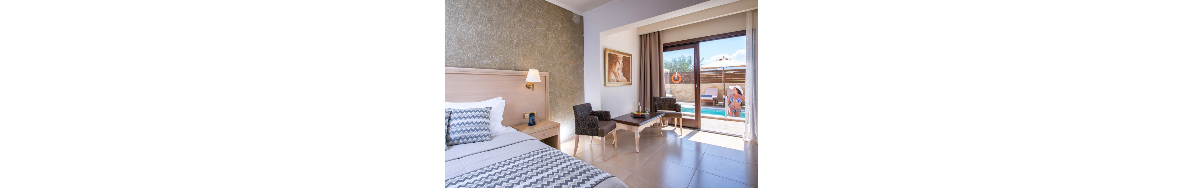 A contemporary hotel room features a neatly made bed with patterned pillows and a wooden headboard. The space includes a sitting area with two chairs and a table, and a large window opens to a private pool where a woman is relaxing outdoors. Sunlight fills the room, highlighting the neutral palette and inviting atmosphere. Visible artwork and decor add a touch of elegance.