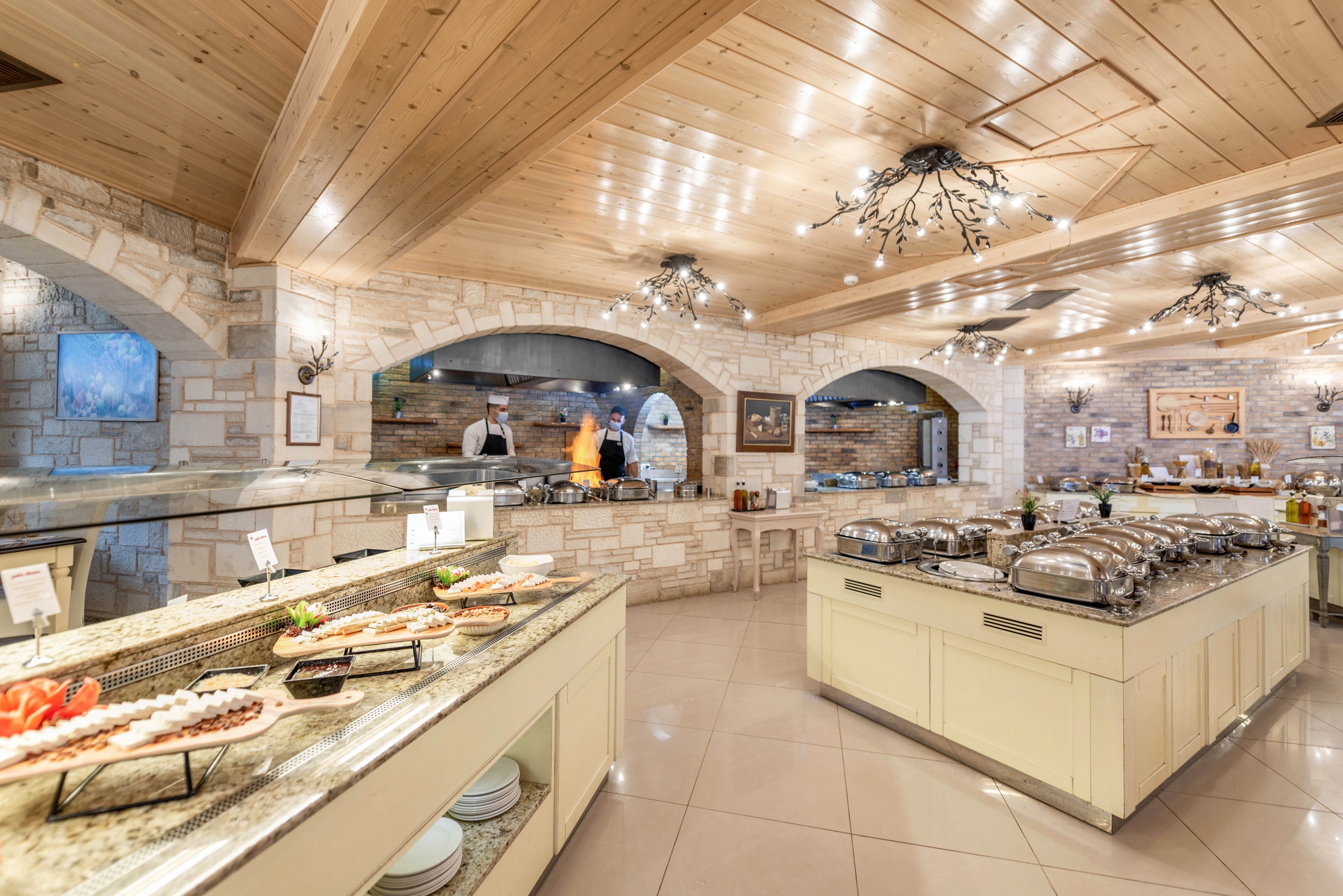 The image shows a spacious buffet restaurant with chefs preparing food behind a counter. The setting features stone walls, wooden ceilings, and elegant lighting fixtures, creating a warm and inviting atmosphere. Multiple food stations display a variety of dishes, including salads and hot entrees, with stainless steel serving trays. The overall mood is bright and welcoming, suitable for casual dining or events.