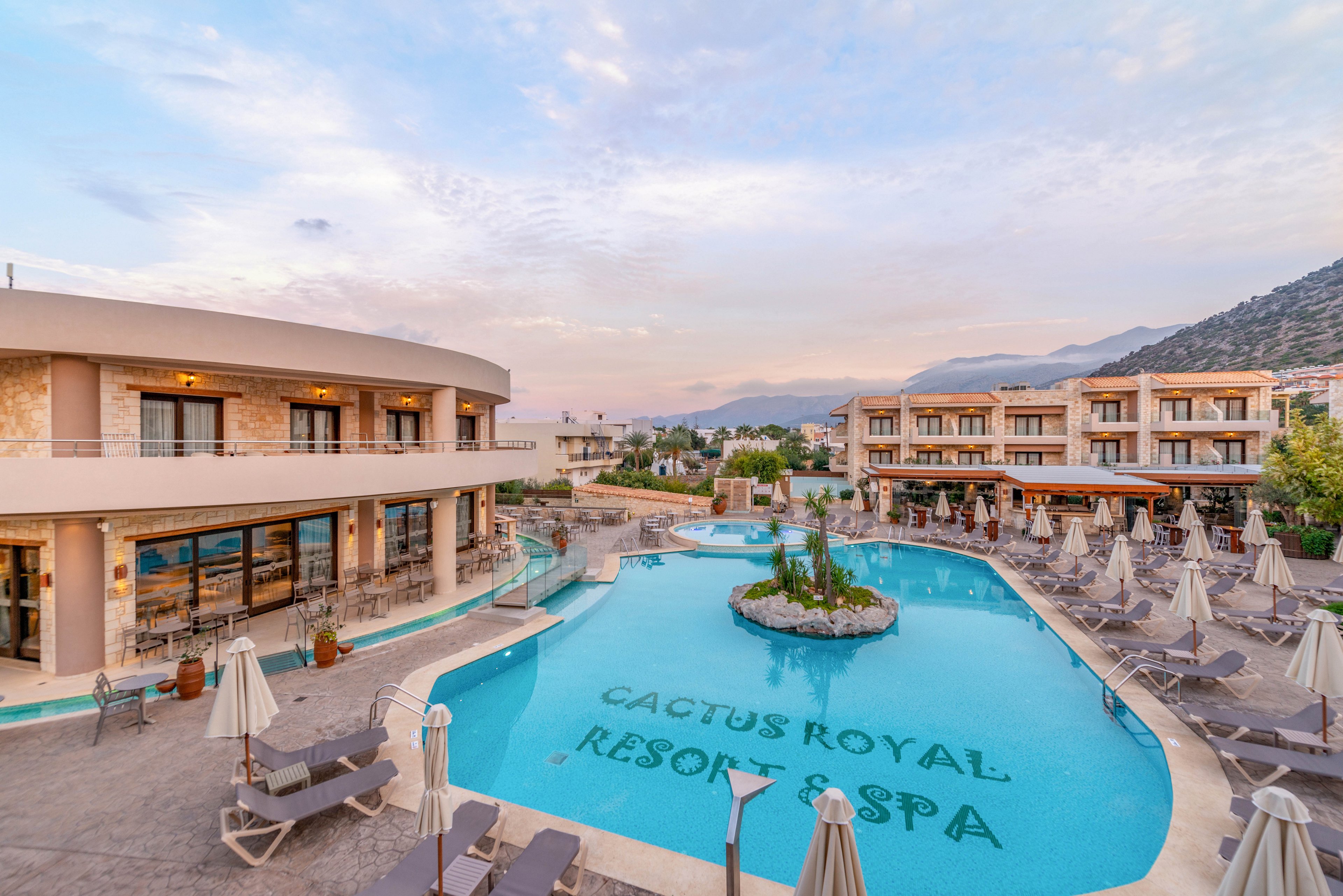 The image features the Cactus Royal Resort Spa, with its name clearly visible in large letters on the bottom of the outdoor swimming pool. The setting is a tranquil resort environment, with modern buildings, numerous poolside loungers, and umbrellas arranged around the pool. The sky is softly colored with pastel hues, suggesting early evening or sunset, and the overall mood is calm and inviting. No people are visible in the scene.