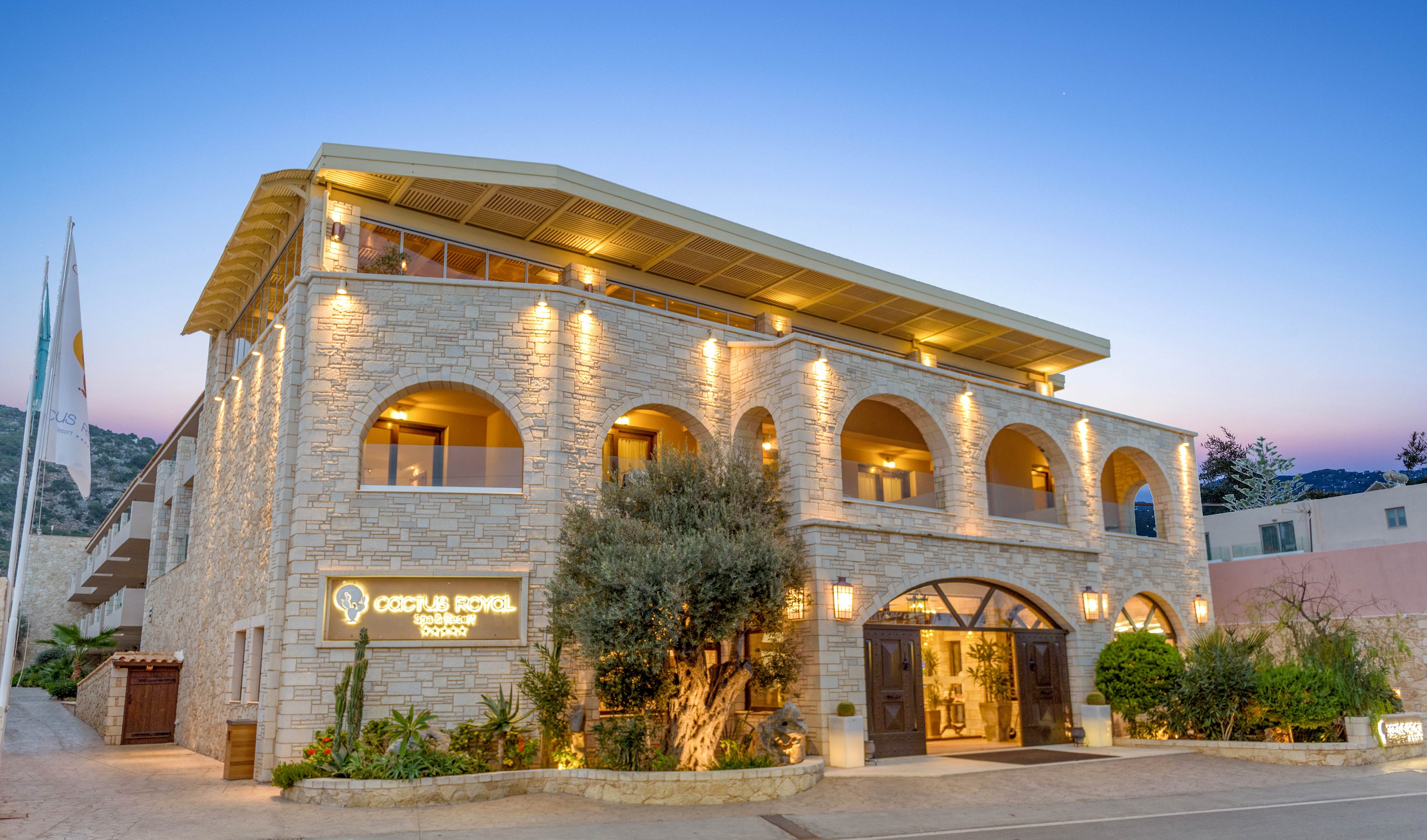 The image features the illuminated facade of the Golden Royal hotel, constructed with light stone and arched windows. The setting is outdoors during dusk, with warm lighting highlighting architectural details and a clear sky in the background. Visible signage reads 'Golden Royal,' and the building is surrounded by greenery and a paved street. The overall mood is inviting and elegant, with a Mediterranean style.
