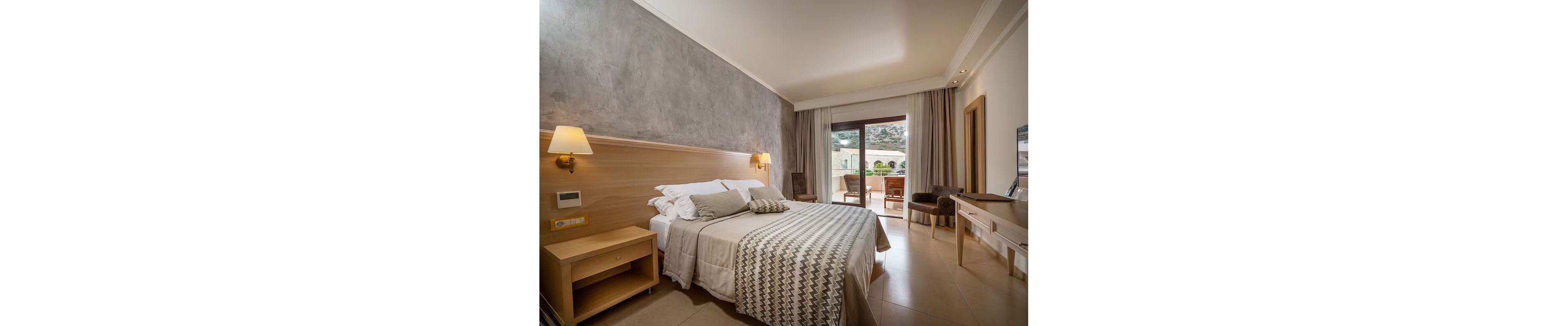 A contemporary hotel room features a large double bed with patterned bedding and multiple pillows. The room is decorated in neutral tones with a textured accent wall and minimalist wooden furniture. A sliding glass door opens to a private balcony with outdoor seating, offering a scenic view of mountains and greenery. Soft lighting and natural daylight create a calm and inviting atmosphere. No visible text or numeric values are present in the image.