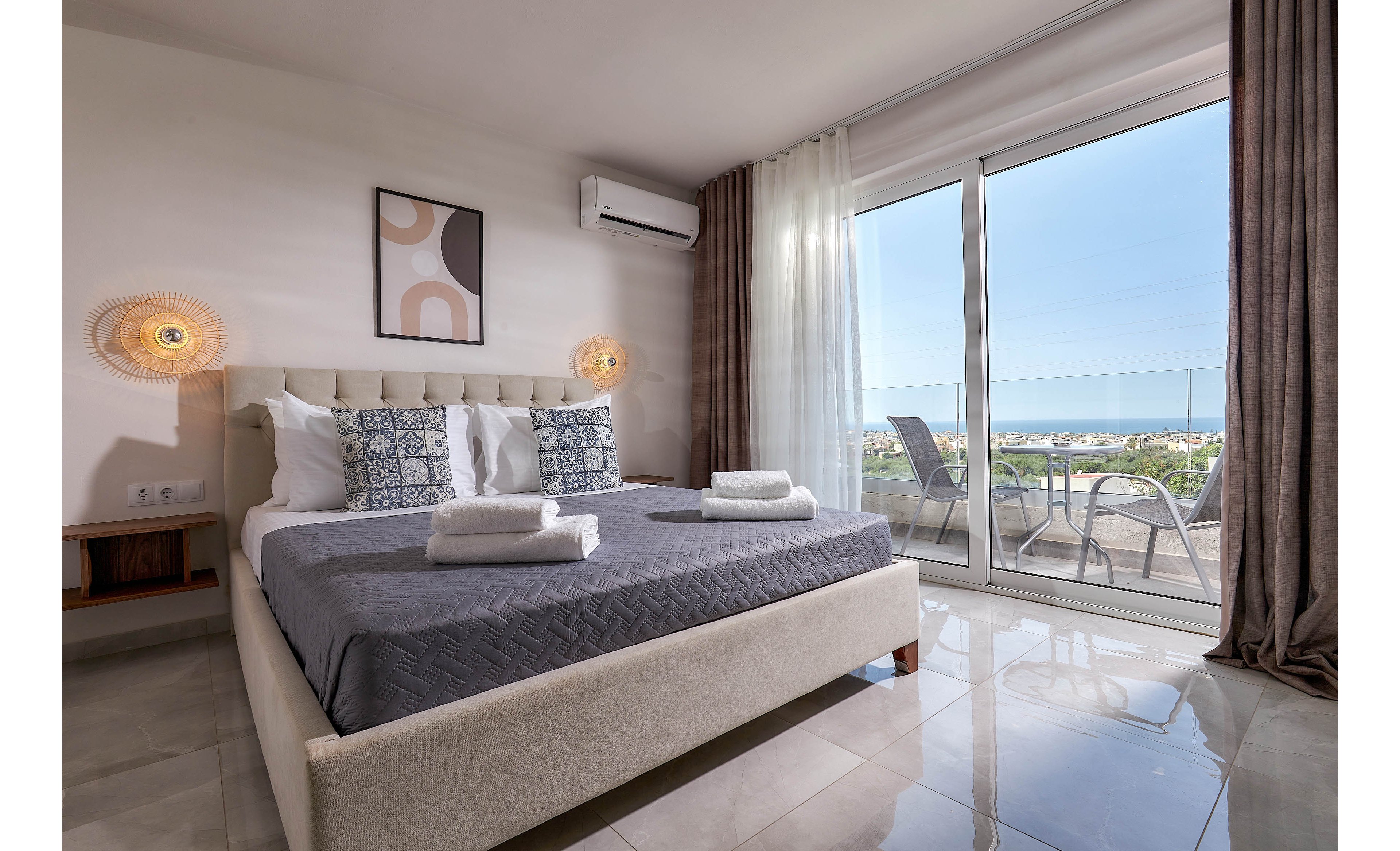 A contemporary hotel bedroom features a large bed with patterned pillows and neatly folded towels. The room is bright, with natural light streaming through floor-to-ceiling glass doors that open to a balcony with outdoor seating. Minimalist decor, neutral tones, and a wall-mounted air conditioner create a calm, inviting atmosphere. The balcony overlooks a lush landscape under a clear sky.