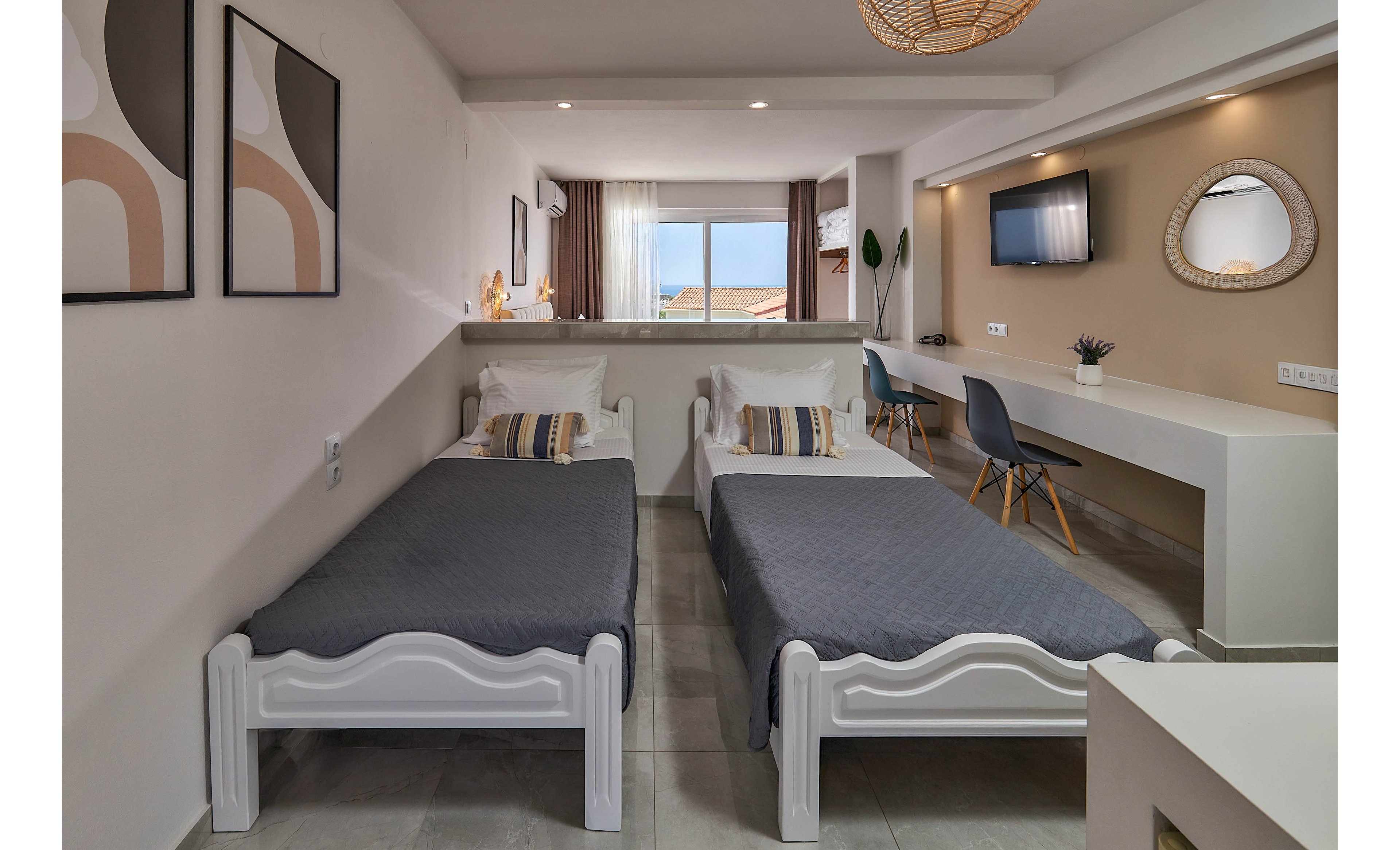 A contemporary hotel room features two single beds with gray bedding and striped pillows. The space includes a long white desk, a wall-mounted television, and a decorative round mirror. Large windows allow natural light to fill the room, highlighting minimalist decor and neutral tones. Abstract artwork and a woven ceiling lamp add visual interest to the setting.