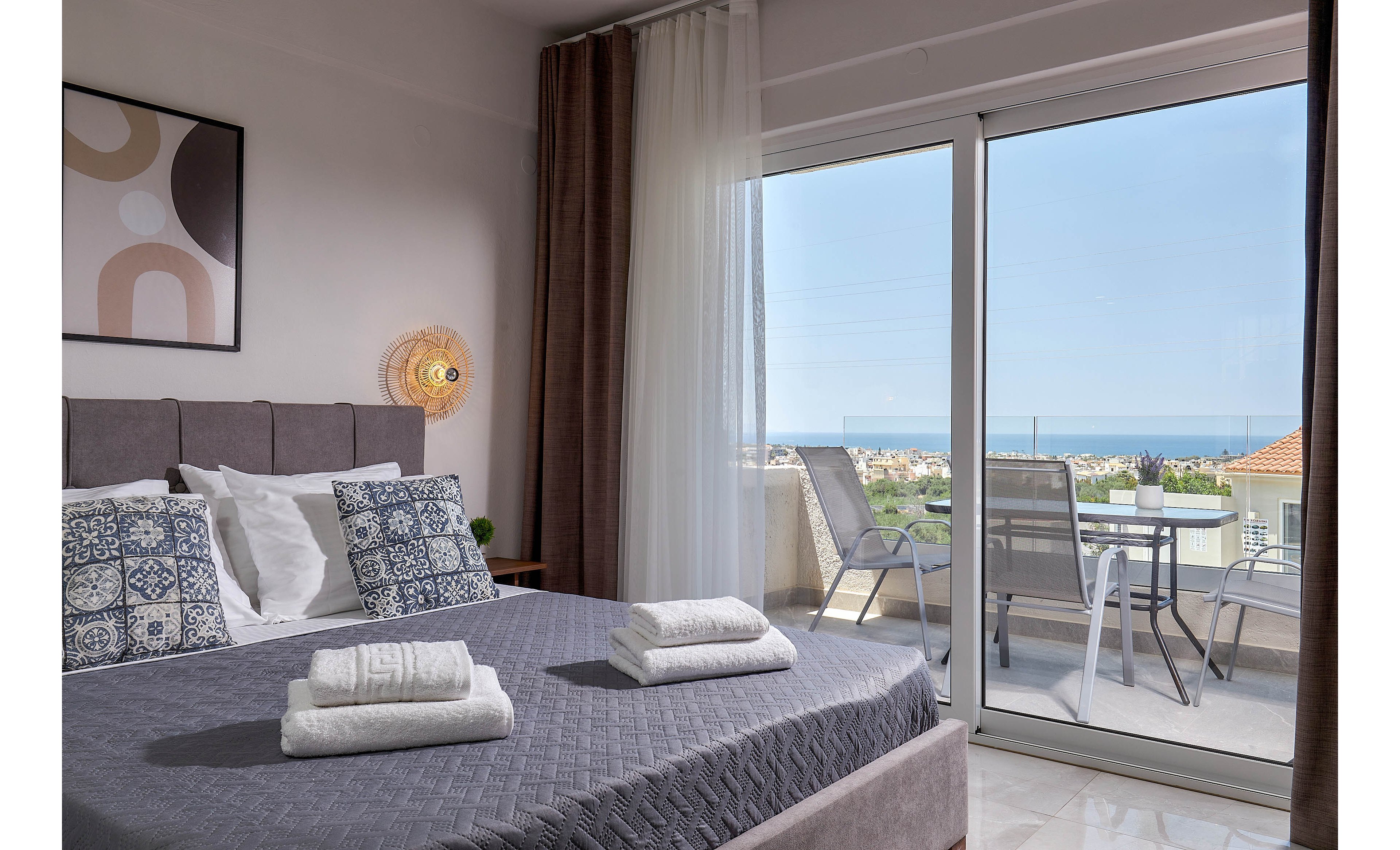 A contemporary bedroom features a neatly made bed with patterned pillows and folded towels. The room opens onto a balcony with glass doors, offering a scenic view of the ocean and rooftops. Neutral tones and natural light create a calm, inviting atmosphere, with modern wall art and outdoor seating visible.