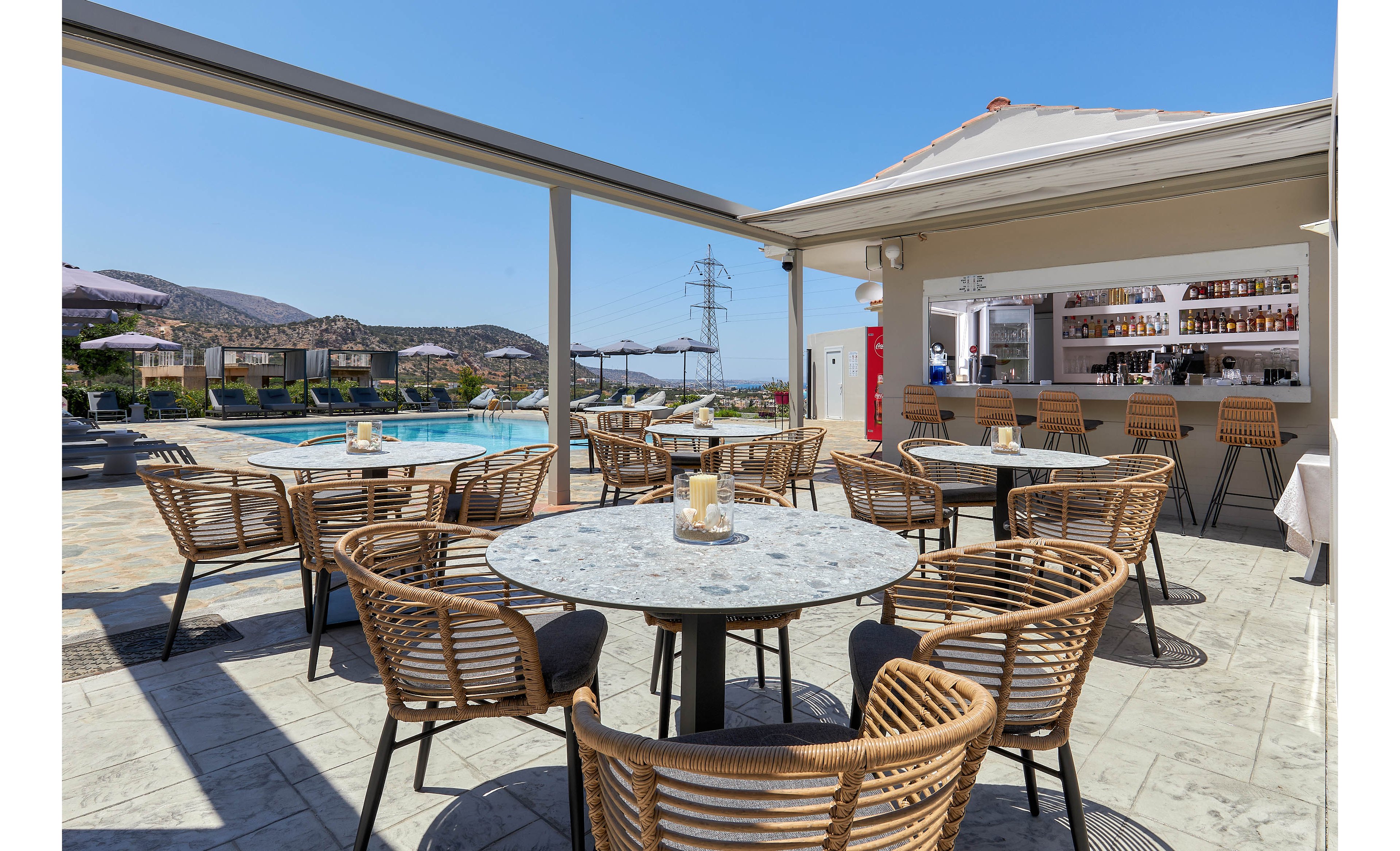 A stylish outdoor bar area features round marble tables and wicker chairs arranged under a shaded pergola. The setting overlooks a swimming pool with sun loungers and a scenic mountain backdrop. The bar is stocked with bottles and glassware, and the atmosphere is bright and inviting with clear blue skies. No visible people are present, and the overall palette is neutral with natural textures.