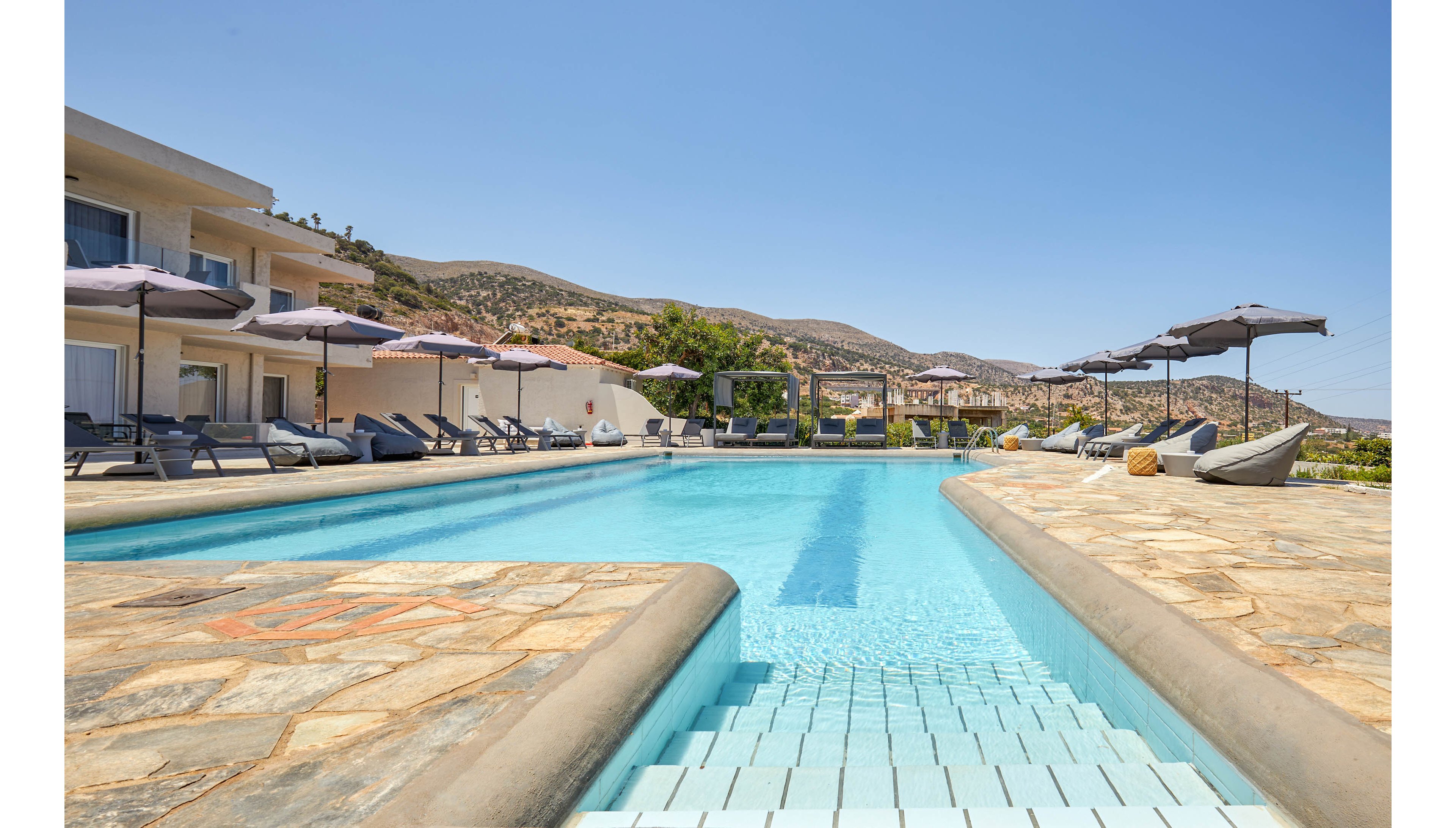 A spacious outdoor swimming pool is the central feature of a modern resort, surrounded by sun loungers and large umbrellas. The setting is bright and sunny, with clear blue skies and distant hills in the background. The pool area is paved with stone tiles, creating a relaxed and inviting atmosphere. No visible text or numbers are present in the image.
