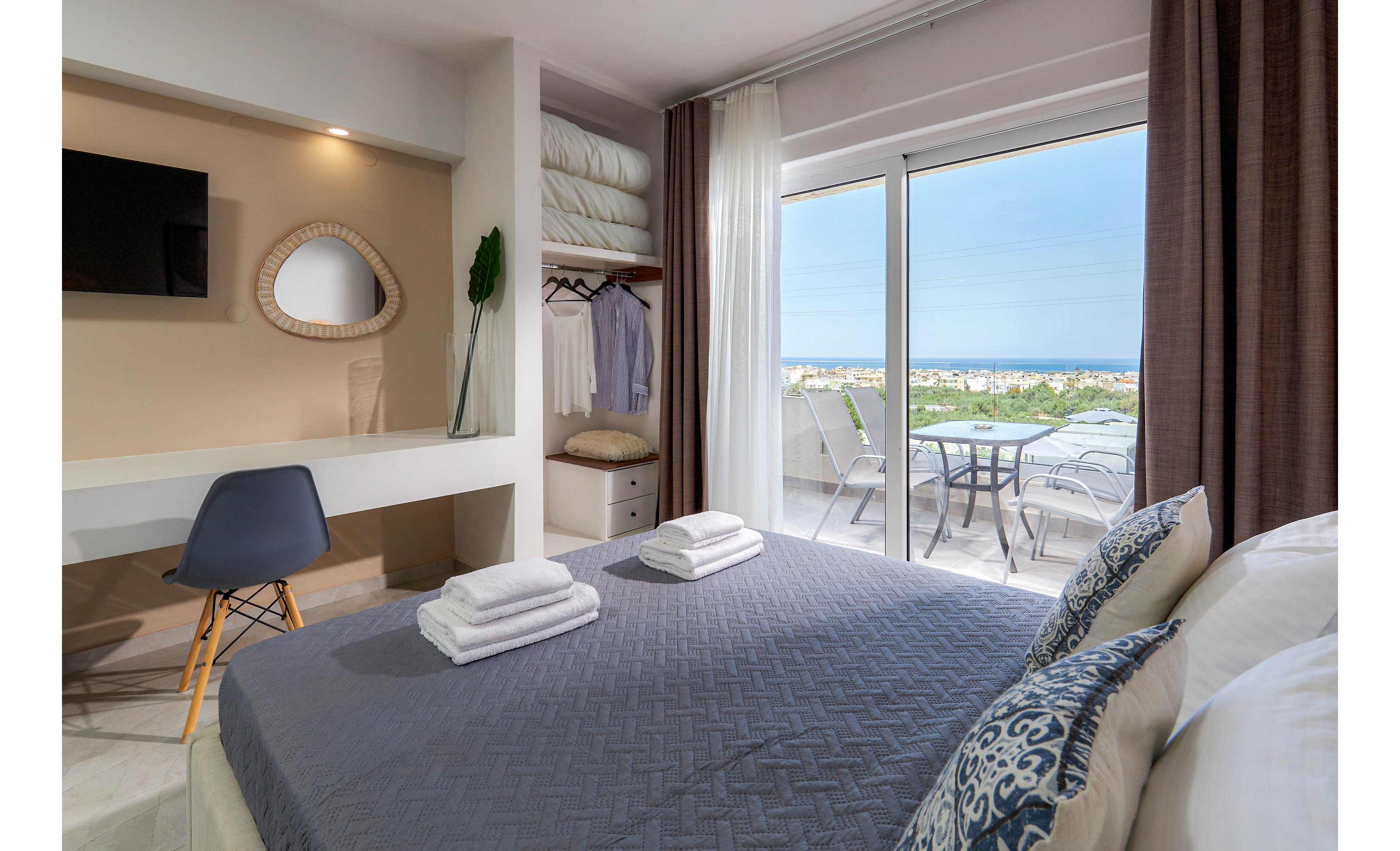 A contemporary hotel room features a neatly made bed with folded towels, a small workspace with a chair, and an open wardrobe. Large glass doors open to a balcony with outdoor seating, overlooking a lush landscape. The room is decorated in neutral tones with blue accents, creating a calm and inviting atmosphere.