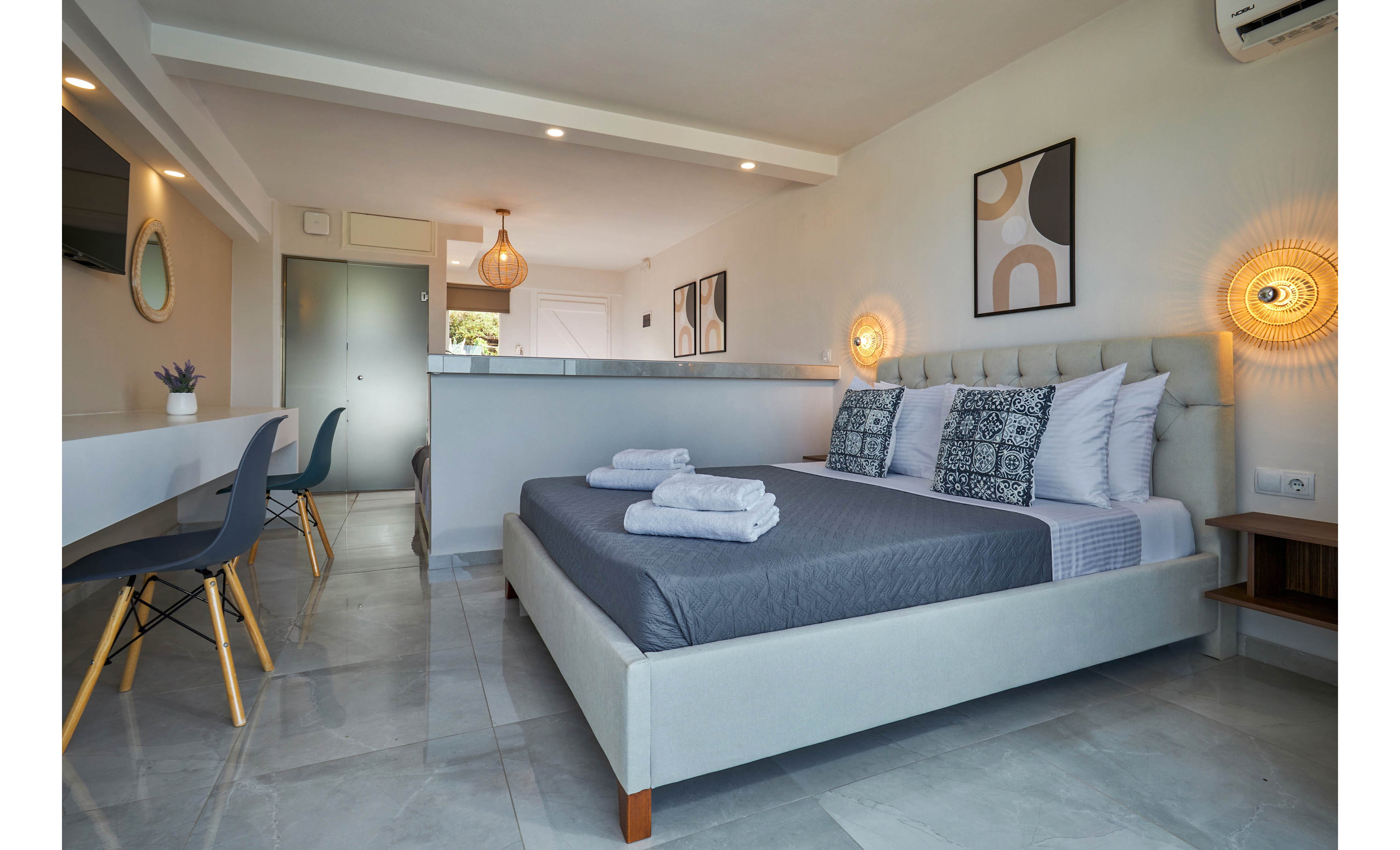 A contemporary bedroom features a neatly made bed with grey bedding and patterned pillows, complemented by folded towels on top. The room includes a workspace with chairs, wall art, and stylish lighting fixtures, creating a clean and inviting atmosphere. The palette is neutral with soft greys and whites, and the space is well-lit with natural and artificial light. No visible text or numbers are present in the image.