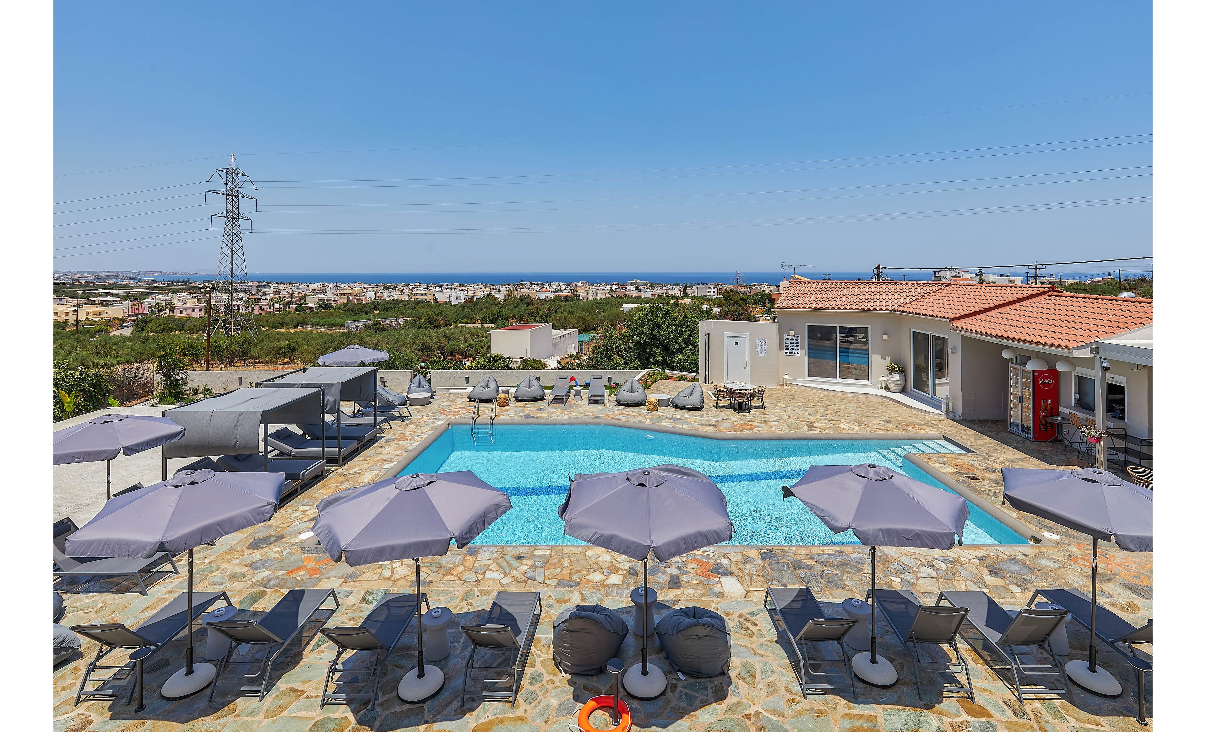 A spacious outdoor swimming pool is surrounded by multiple sun loungers and large umbrellas, creating a relaxing resort atmosphere. The pool area is set against a backdrop of a clear blue sky and distant cityscape, with a modern pool house and shaded seating visible. The stone-tiled deck and bright sunlight evoke a summery, tranquil mood. No visible text or numbers are present in the image.