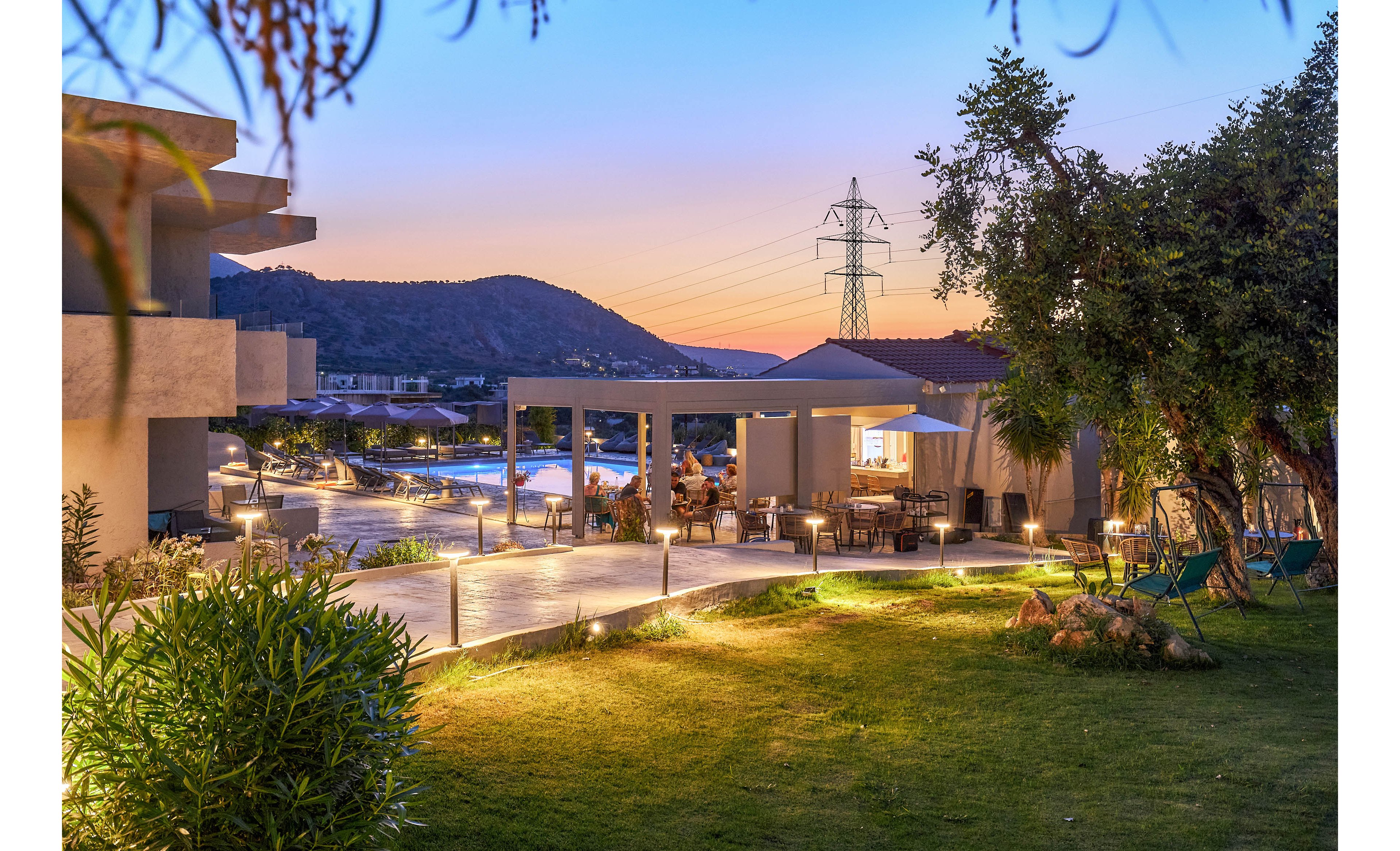 A contemporary resort terrace overlooks a glowing swimming pool at sunset. The outdoor seating area is softly illuminated, with guests enjoying the tranquil evening ambiance. Lush greenery surrounds the space, and distant hills and power lines are visible against the colorful sky. The scene conveys relaxation and leisure in a scenic, modern setting.