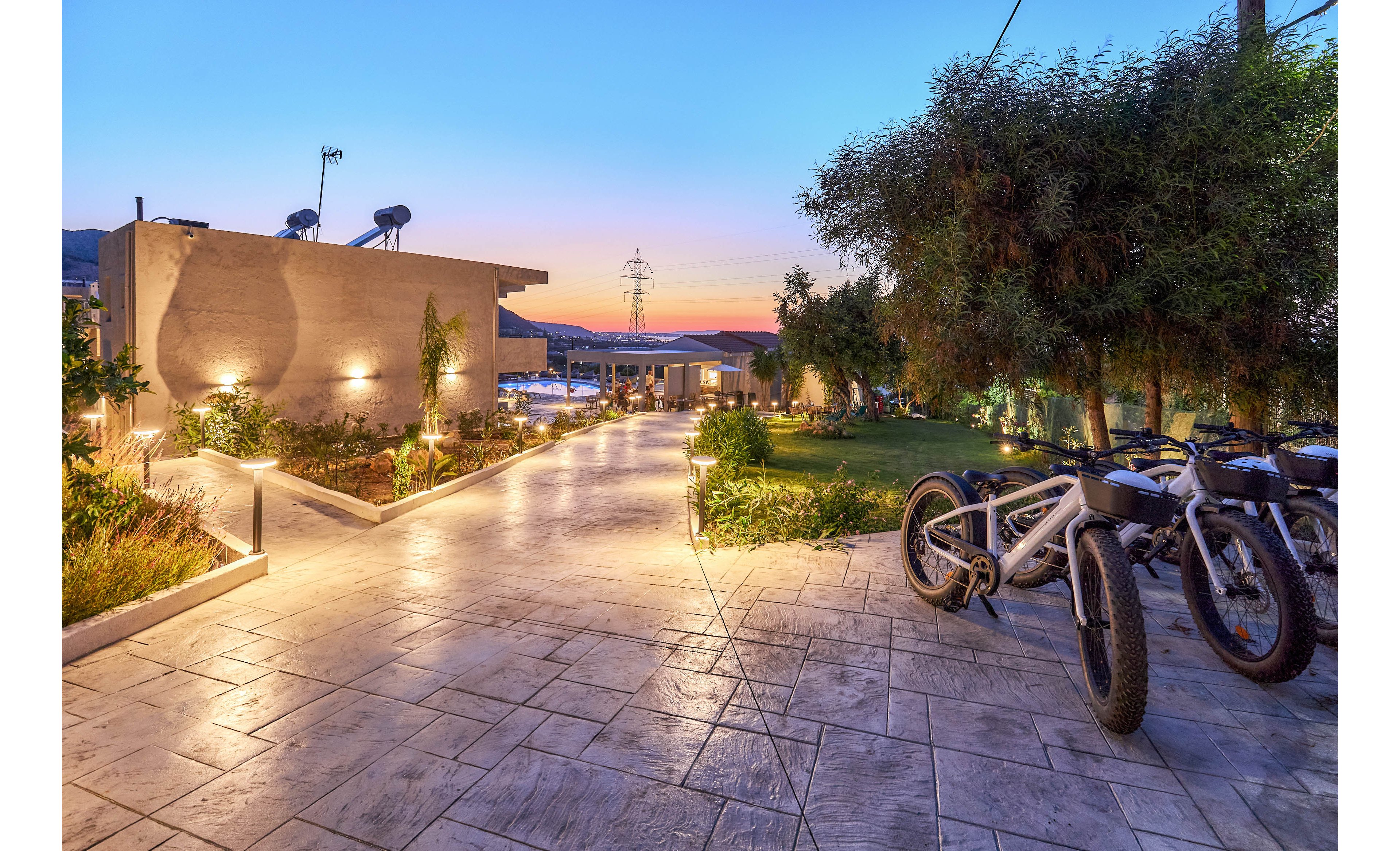 A contemporary villa entrance is illuminated by warm pathway lights during sunset. Two bicycles are parked on the right side, suggesting outdoor activity or guest amenities. The paved walkway leads to the main building, surrounded by landscaped greenery and distant mountain views. The tranquil setting evokes a relaxed, inviting atmosphere.