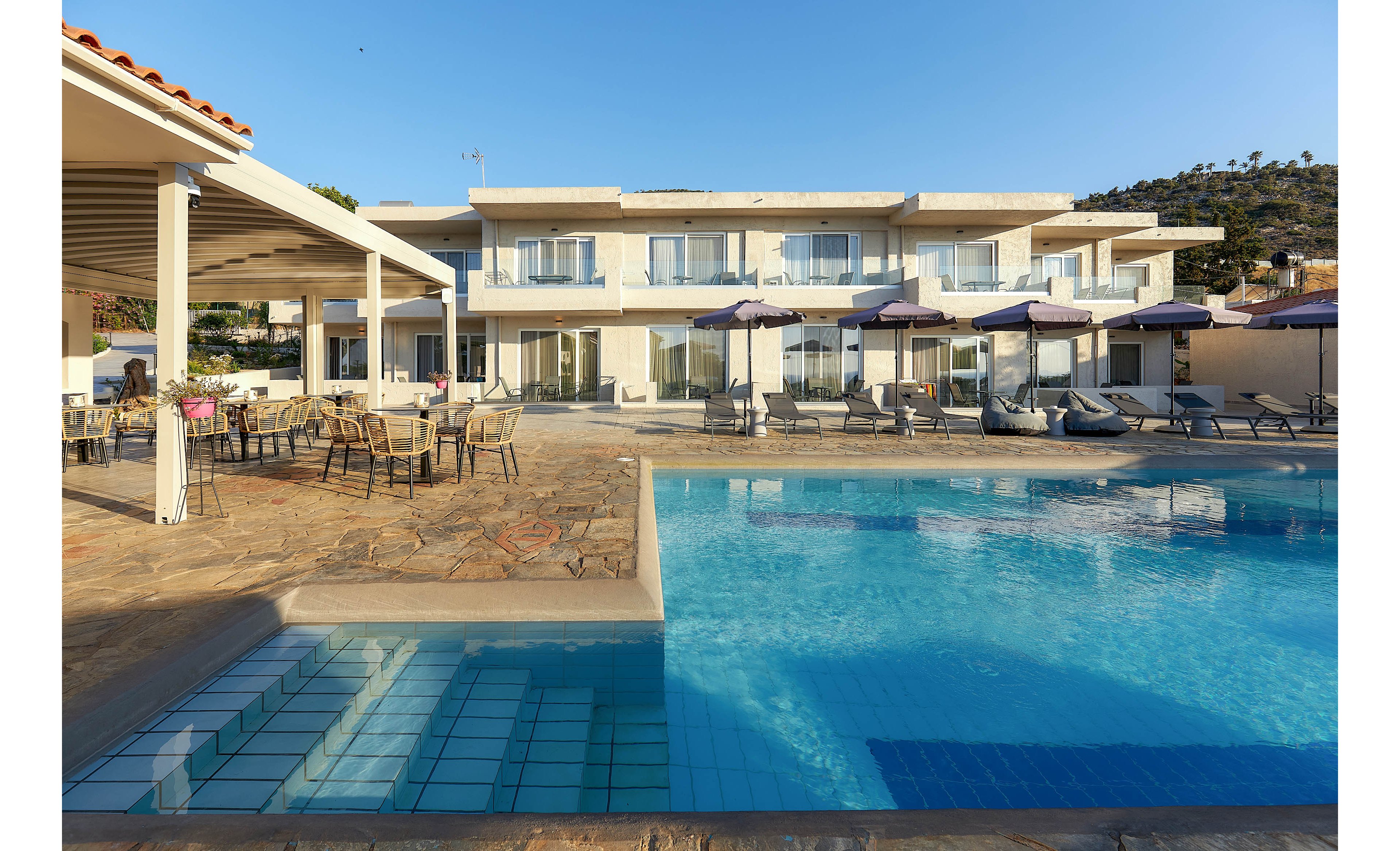 A contemporary hotel building features a spacious outdoor swimming pool in the foreground. Sun loungers and umbrellas line the poolside, while a shaded patio area with tables and chairs offers a relaxing spot for guests. The setting is bright and sunny, with clear blue skies and a tranquil atmosphere. No visible text or numbers are present in the image.