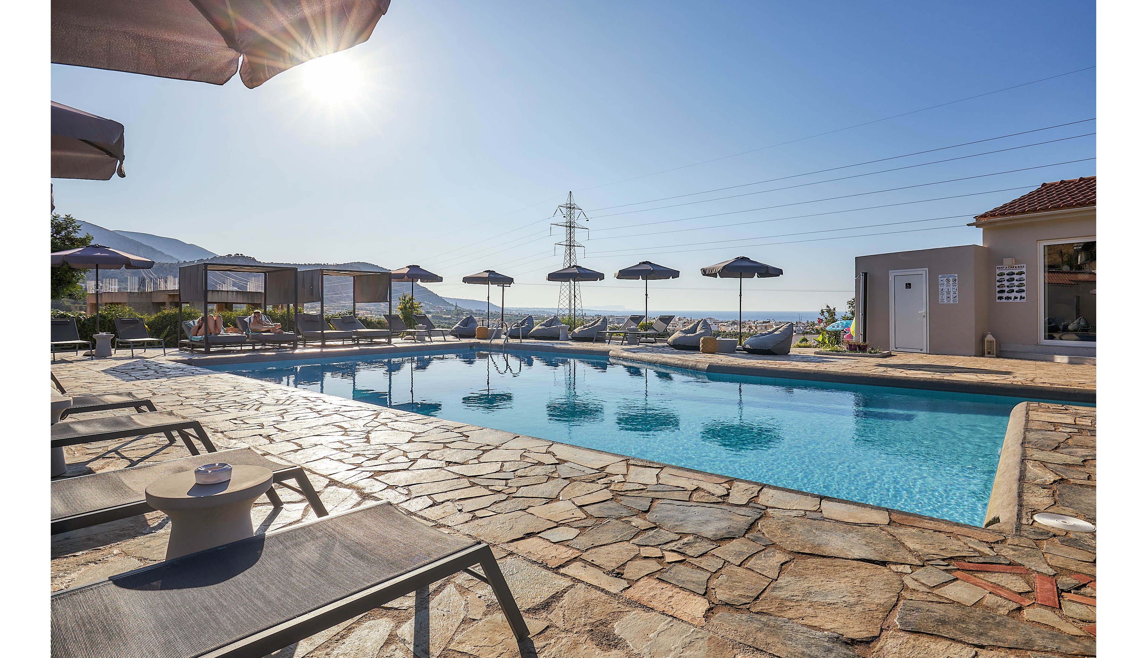 A tranquil outdoor swimming pool is surrounded by stone paving and several lounge chairs under black umbrellas. The setting is sunlit, with clear blue skies and a scenic view of distant hills and the sea. The atmosphere is calm and inviting, ideal for relaxation or vacation. No visible people or text are present in the image.