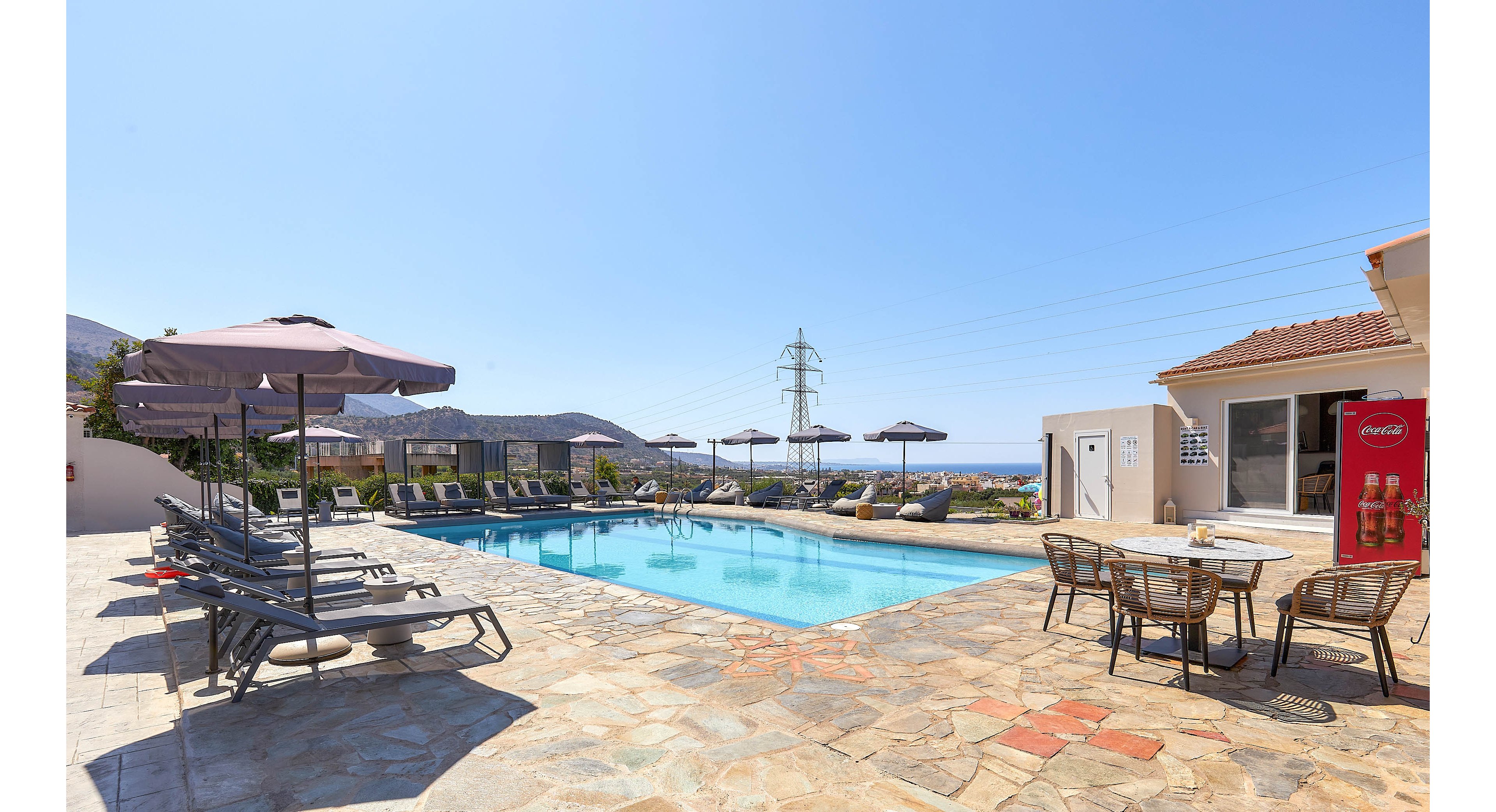 A bright outdoor swimming pool is surrounded by lounge chairs and large umbrellas, set on a stone patio. The scene includes a table with chairs and a visible beverage vending machine with the word 'CocaCola' clearly displayed. The setting is tranquil, with clear blue skies and distant hills, evoking a relaxed vacation atmosphere.