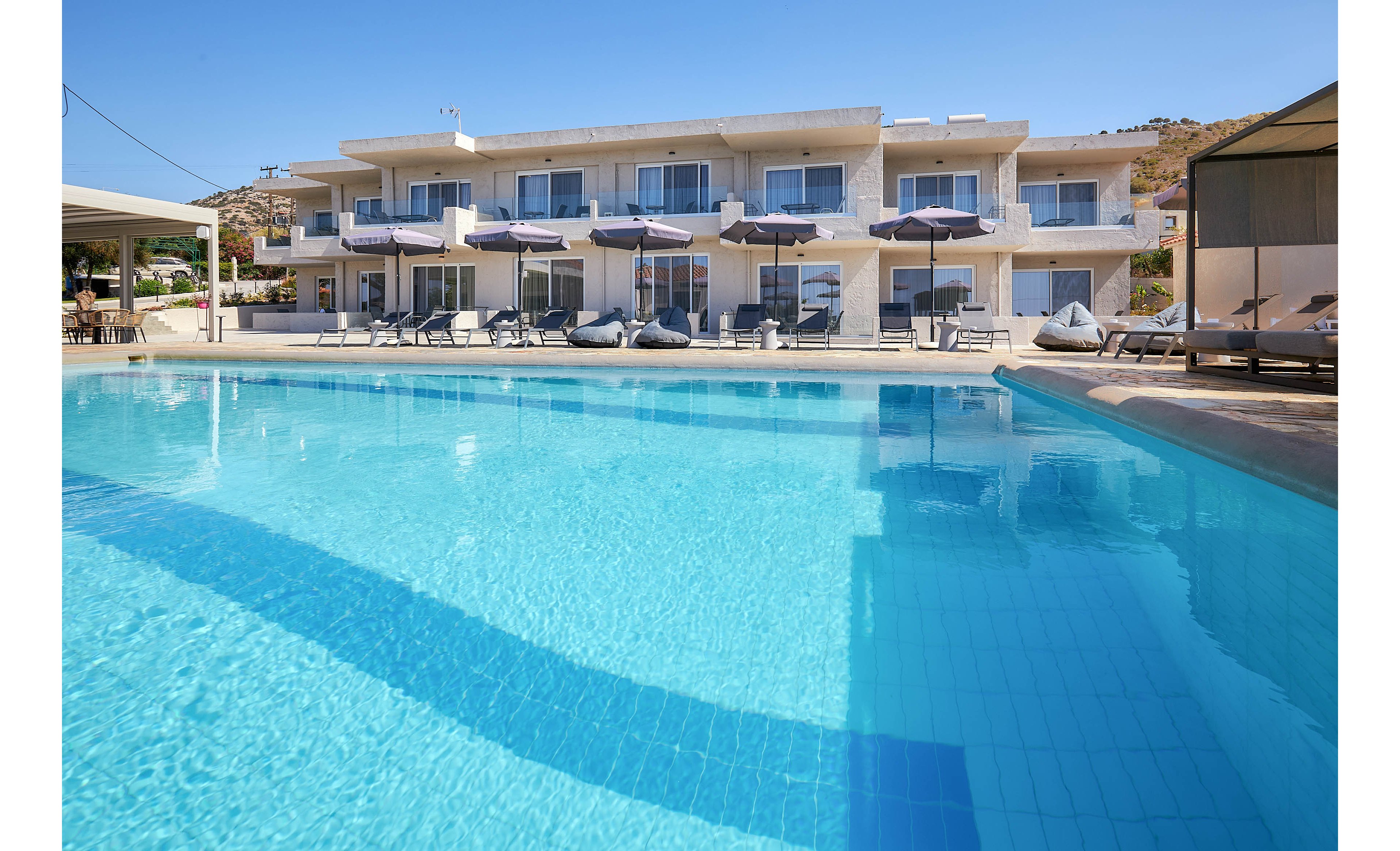 A contemporary hotel building features multiple balconies overlooking a spacious outdoor swimming pool. Sun loungers and umbrellas are arranged around the poolside, creating a relaxing vacation atmosphere. The scene is set in bright daylight with clear blue water and minimalistic architecture. No visible text or numbers are present in the image.
