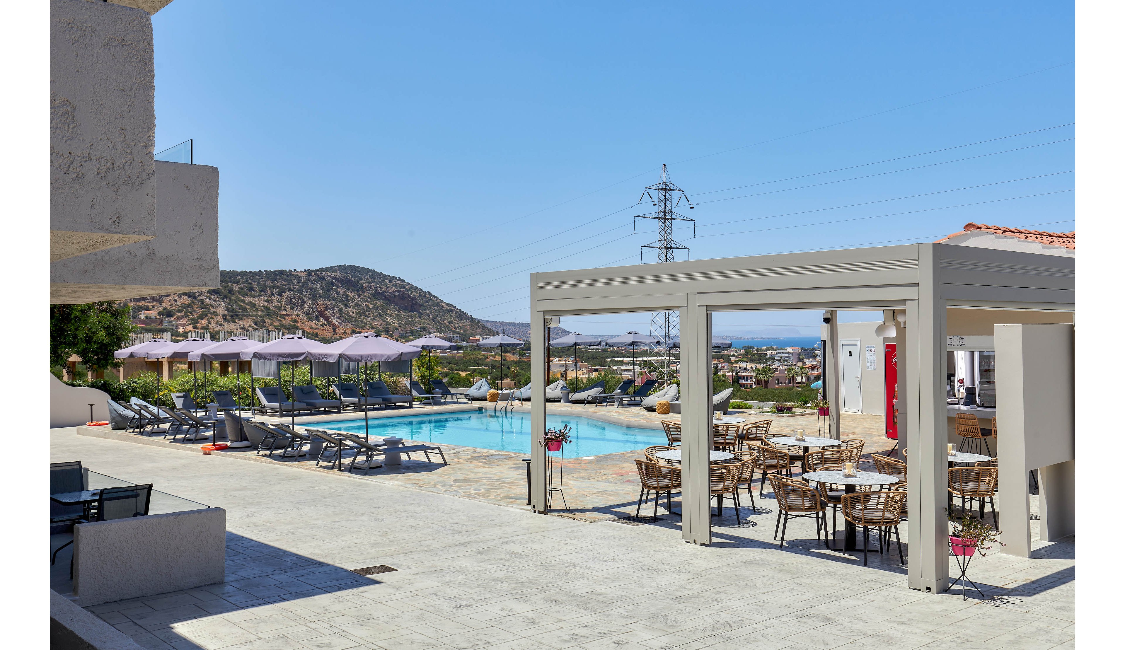 A spacious outdoor pool area features lounge chairs and shaded tables under a pergola. The setting overlooks distant hills and a clear blue sky, with a power line visible in the background. The pool is surrounded by sun umbrellas and offers a relaxing atmosphere for guests. No visible text or numbers are present in the image.
