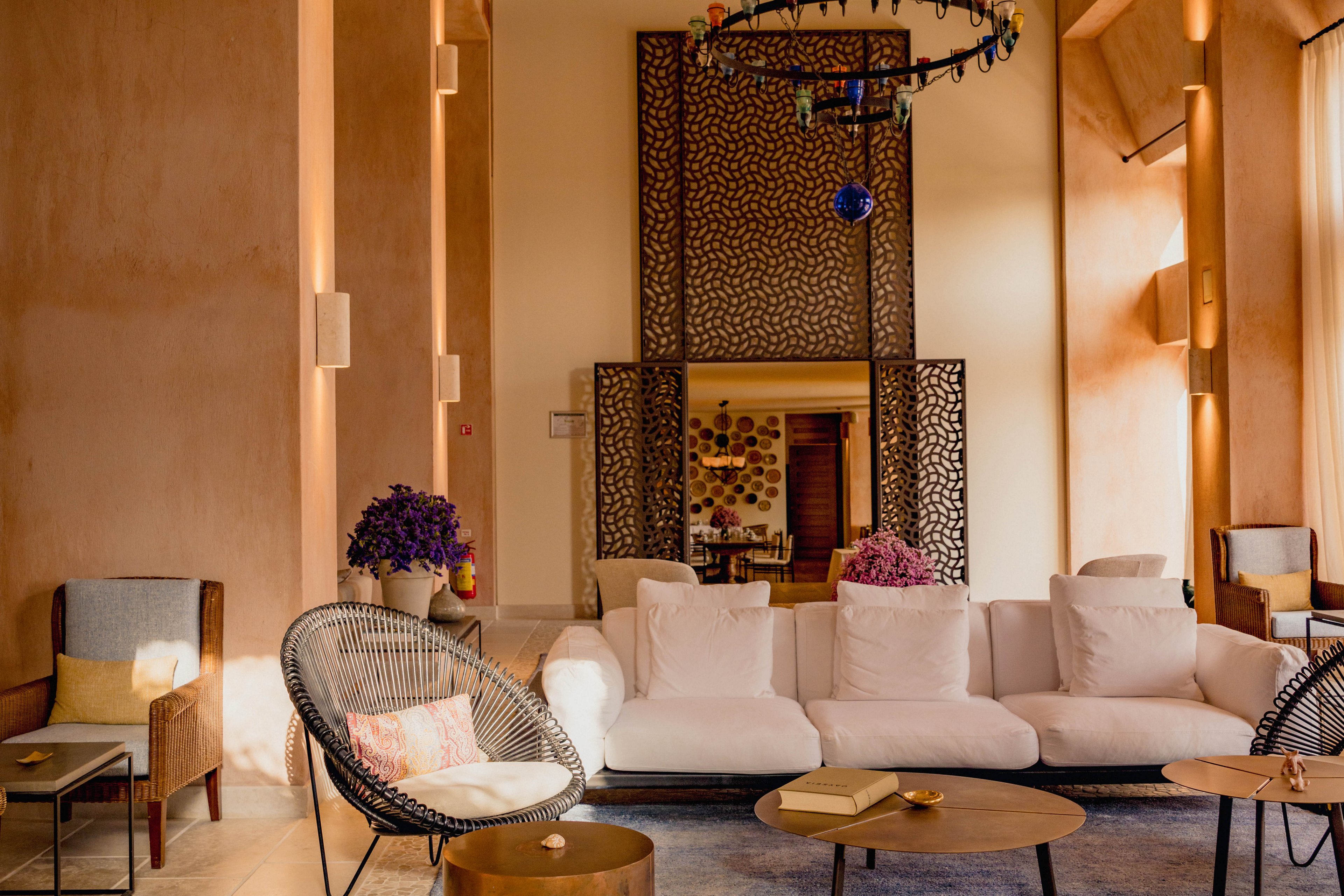 A stylish lounge area features a white sofa and modern chairs arranged around a low table on a blue rug. The space has tall walls with warm beige tones and exposed wooden ceiling beams. Decorative screens and a colorful hanging chandelier add visual interest in the background. Soft natural light enters from large windows, creating a cozy and inviting atmosphere.