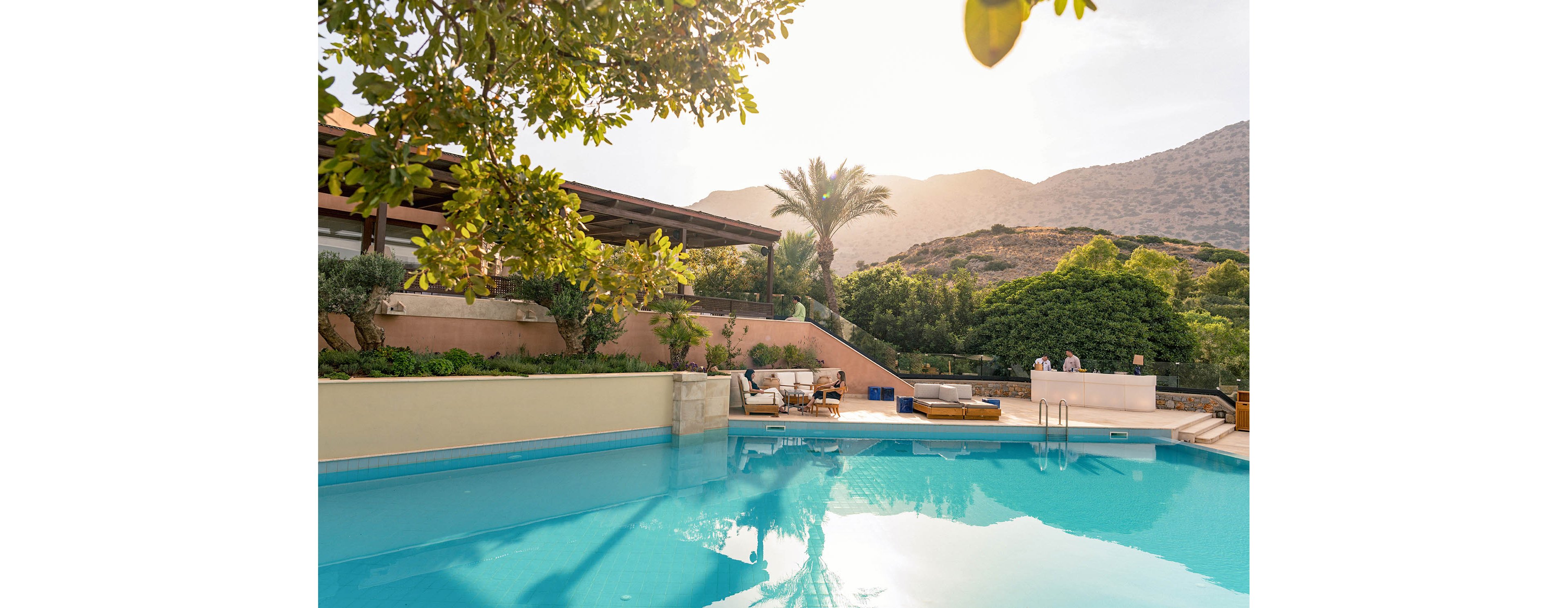 An inviting outdoor swimming pool reflects the warm light of a setting sun at a luxury resort. Lounge chairs and seating areas line the poolside terrace beside a modern villa-style building. Lush greenery, palm trees, and distant hills create a tranquil natural backdrop. Two people stand near the pool edge, enjoying the peaceful evening atmosphere.